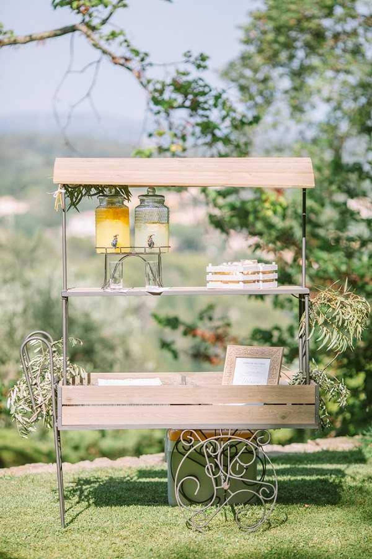 A detail shot of an outdoor cocktail hour or welcome drink station set up on a lawn with a panoramic countryside view in the background. The display unit is a vintage-style metal and light wood three-tier cart with decorative scrollwork at the base, styled in a rustic-chic aesthetic. The top shelf holds two glass drink dispensers — one filled with a yellow citrus beverage and one with clear water — alongside stacked glasses, while the middle shelf displays a small wooden crate of individual packaged favors or snacks. The bottom shelf holds folded white napkins and a framed sign, with dried botanical elements hanging from the sides of the cart.