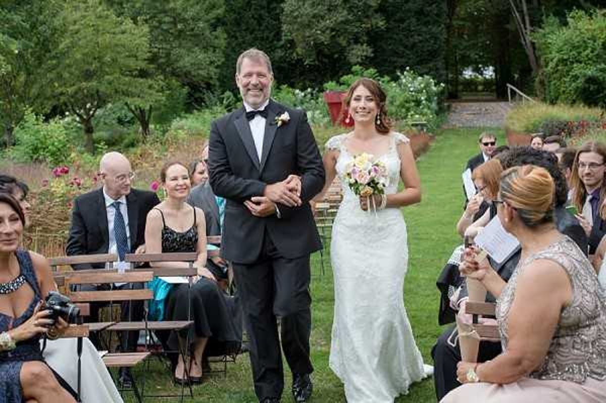 An outdoor garden wedding ceremony showing a bride being walked down the aisle by a male companion, presumed to be her father. The bride wears a fitted white lace gown with cap sleeves and carries a bouquet of pale pink and white blooms with greenery. Her escort is dressed in a black tuxedo with a bow tie and a small boutonniere. Seated guests line both sides of the wooden folding chair aisle, dressed in formal attire including a woman in a black dress and another in a teal outfit. An officiant in a silver beaded dress stands to the right holding papers. The setting is a lush garden with mature trees and shrubs in the background. Wide shot taken from near the front of the aisle looking back toward the couple.