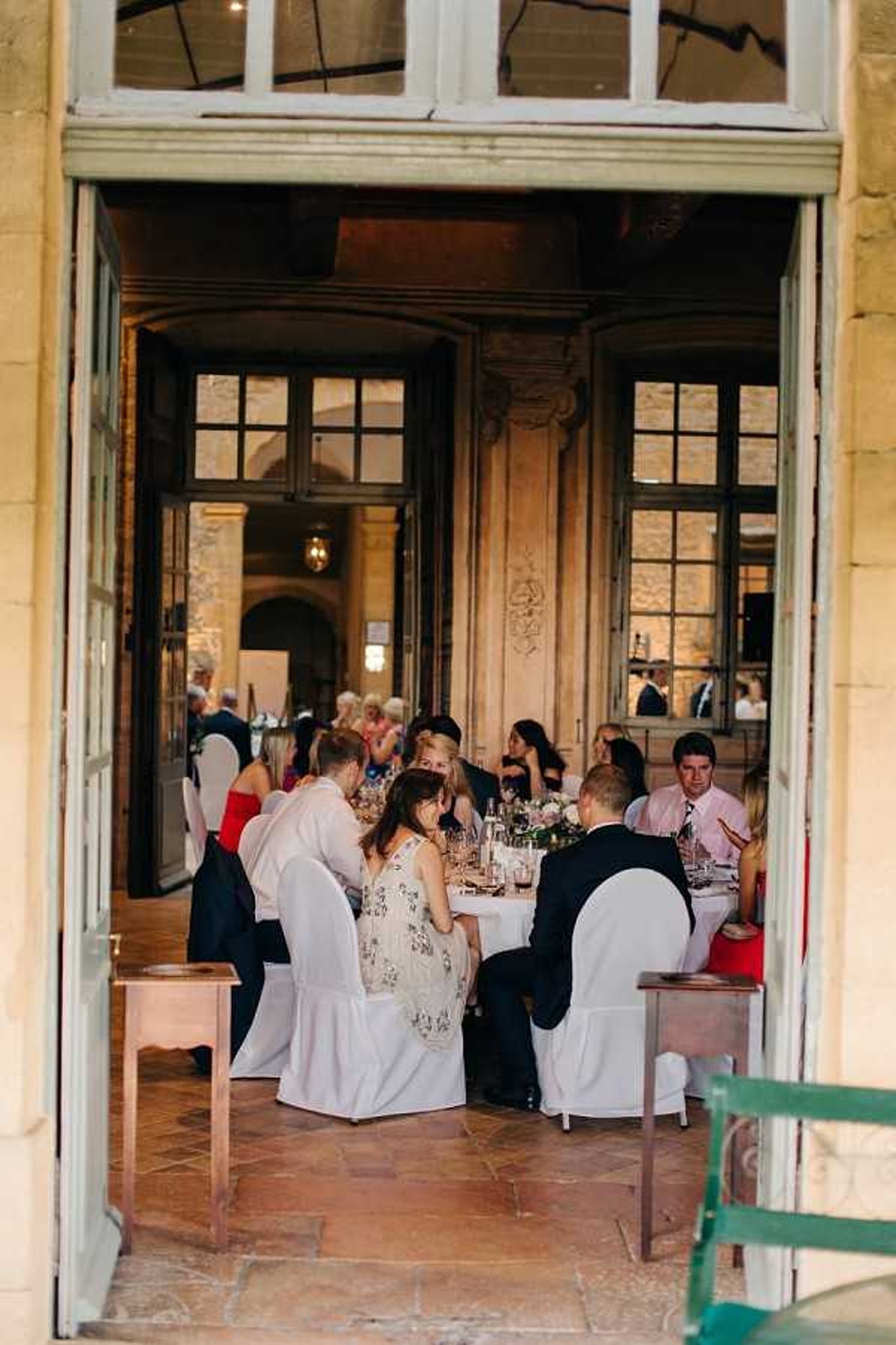 Wedding reception dinner scene photographed from outside through large open French doors, looking into a warmly lit interior room of what appears to be a historic chateau or manor. Approximately 15–20 guests are seated around round tables covered with white linens, with glassware and low floral centerpieces visible on the tables. Guests are dressed in formal attire including black suits, a red dress, a floral patterned dress, and a pink shirt with tie. The interior walls feature ornate carved wood paneling and large multi-pane windows, with warm ambient lighting creating a golden tone. The shot is taken from a terrace or exterior corridor with terracotta tile flooring, framing the scene through the doorway in a wide, candid composition. Potential venue feature image.