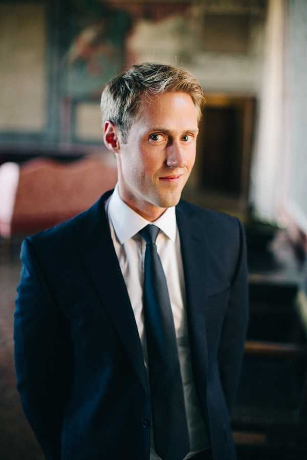 Close-up portrait of a groom standing indoors, wearing a navy suit jacket, white dress shirt, and a dark charcoal tie. He is looking directly at the camera with a slight smile. The background is softly blurred but shows what appears to be an ornate interior room with aged decorative paintings or frescoes on the wall and dark wooden furniture, suggesting a historic venue such as a chateau or manor house. The lighting is natural and slightly warm, coming from a window to the right of frame.
