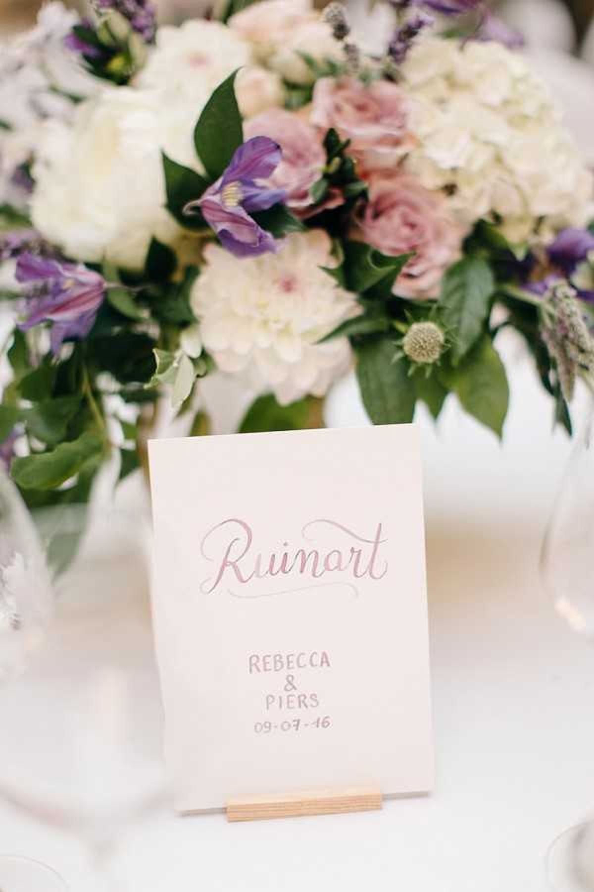 A close-up detail shot of a reception table setting featuring a calligraphy table name card reading 'Ruinart' with the couple's names 'Rebecca & Piers' and date '09.07.16' written in a dusty rose script on cream card stock, displayed in a small wooden holder. Behind the card is a lush floral centerpiece arranged in a white vessel, composed of ivory hydrangeas, blush roses, purple clematis or iris blooms, lavender sprigs, thistle buds, and green foliage. The overall decor palette is soft and romantic, combining cream, blush, and purple tones against white table linens, consistent with a classic or garden-inspired reception styling.