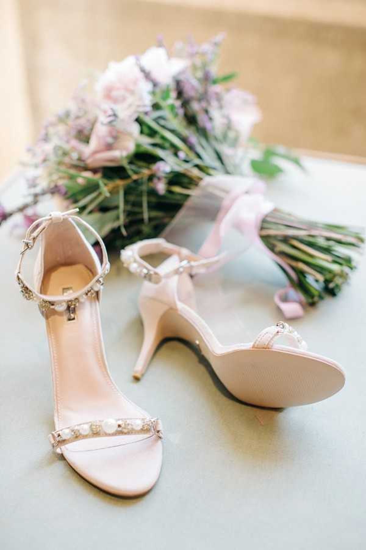 A close-up detail shot of a pair of blush pink strappy heeled sandals adorned with pearl and crystal embellishments on the toe strap and ankle strap, placed on a pale blue surface. In the soft-focus background lies a bridal bouquet composed of blush pink roses, lavender wisteria or statice, and green foliage, tied with a pale pink satin ribbon. The composition is a styled flat-lay arrangement shot from a slightly elevated angle with soft, diffused natural light.