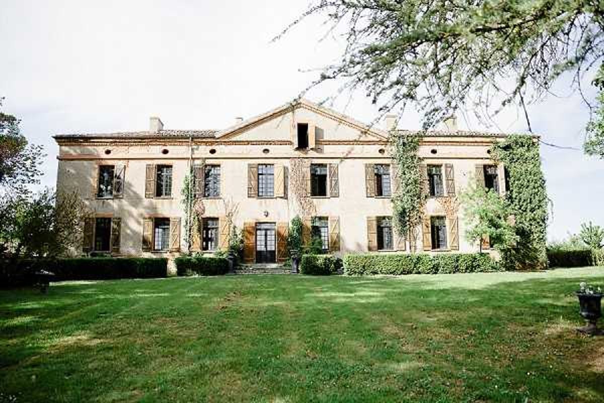 Wide exterior shot of a French country house or small chateau, featuring a symmetrical two-story facade with warm sand-colored render, brown wooden shutters, and a classical pediment above the central entrance. The building is partially covered in climbing ivy on the right side, with trimmed hedging and topiary flanking the front steps. No people are visible in the image; the focus is entirely on the architecture and manicured grounds. Potential venue feature image.