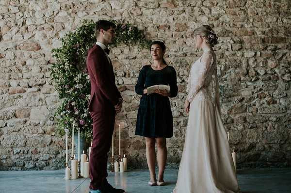 A wedding ceremony is taking place indoors against an exposed stone wall, with the couple facing each other and an officiant standing between them holding a booklet. The groom wears a deep burgundy suit with a bow tie, and the bride wears a long ivory dress with lace long sleeves and her hair pinned up with a floral accent. Behind them is a large circular greenery arch decorated with pink and mauve flowers, flanked by clusters of white pillar candles in varying heights arranged on the concrete floor. The officiant wears a navy blue knee-length dress. The setting has a rustic feel with the raw stone wall backdrop and polished concrete floor. The image is a medium wide shot capturing all three figures full-length.