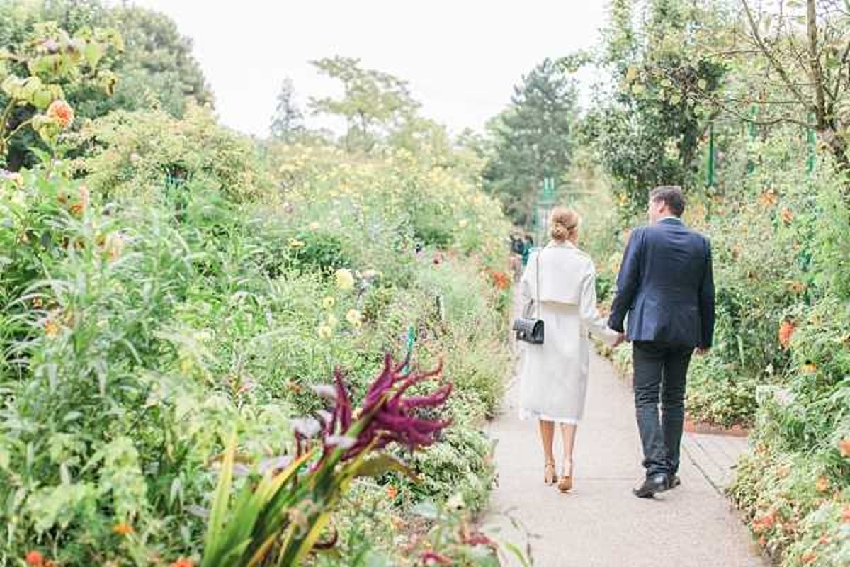 A couple walks hand-in-hand along a garden pathway, photographed from behind in a wide portrait composition. The woman wears a white or cream knee-length dress with a matching white coat and carries a small black crossbody bag, with her hair styled in an updo; the man wears a navy suit with dark trousers. The setting is a lush formal garden with dense, colorful plantings on both sides of the paved path, including dahlias in orange and burgundy tones and a variety of foliage. The overall styling is classic and understated, with a soft, bright natural light giving the image an airy feel.
