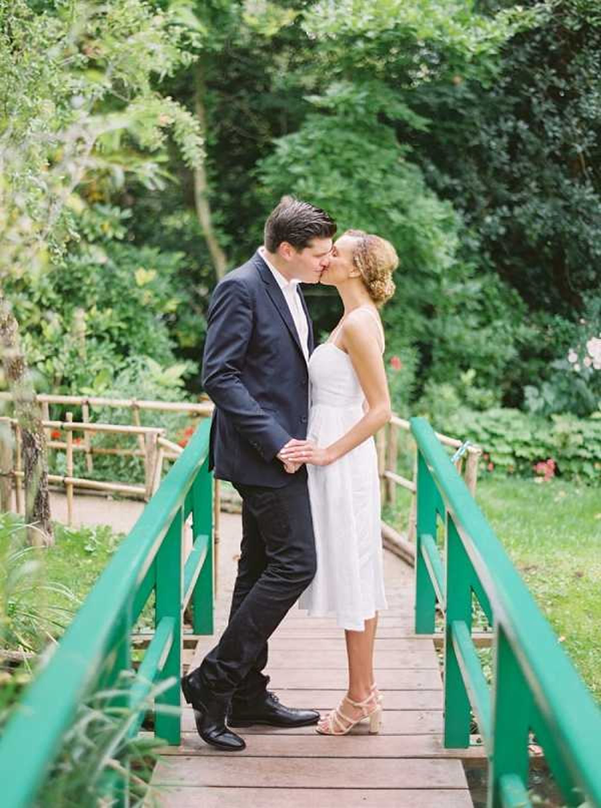 A couple shares a kiss while standing on a small wooden footbridge with green-painted railings, set within a lush garden. The bride wears a knee-length white spaghetti-strap dress and strappy nude heeled sandals, with her hair styled in an updo, while the groom wears a navy suit with black dress shoes. They are holding hands at the center of the bridge, with additional bamboo-railed pathways visible in the background. The shot is a full-length portrait with a romantic, garden-casual style.