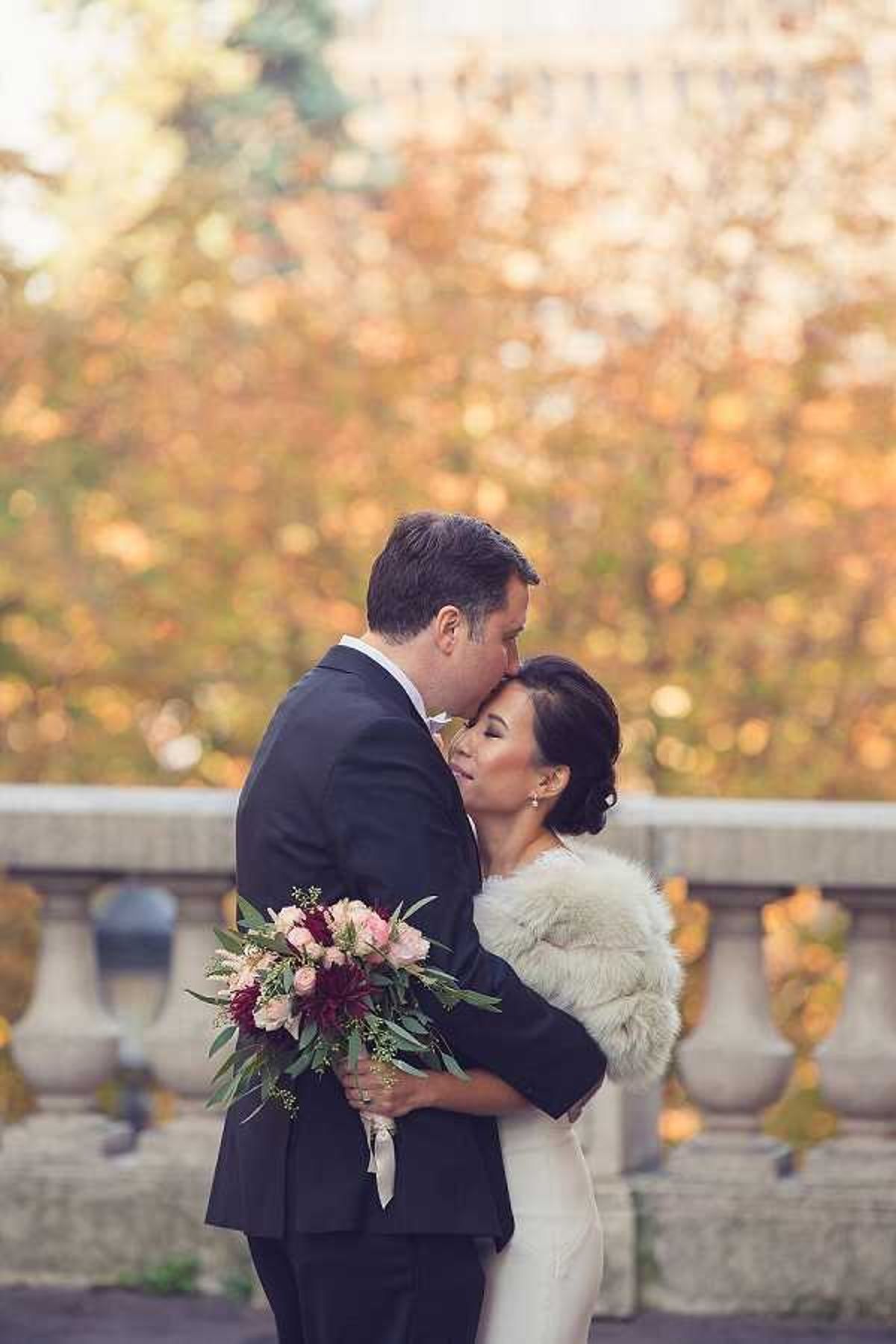 A couple portrait taken outdoors, showing the groom kissing the bride on the temple while embracing her from behind. The groom wears a dark navy suit with a white dress shirt, and the bride wears a white fitted gown layered with a cream fur stole wrap. She carries a loose, garden-style bouquet featuring burgundy dahlias, blush pink roses, and eucalyptus foliage tied with an ivory ribbon. A stone balustrade is visible in the background, with bokeh-blurred autumn foliage in warm amber and gold tones filling the background. The image has a warm, soft color palette suited to a fall wedding. Medium portrait shot with shallow depth of field.
