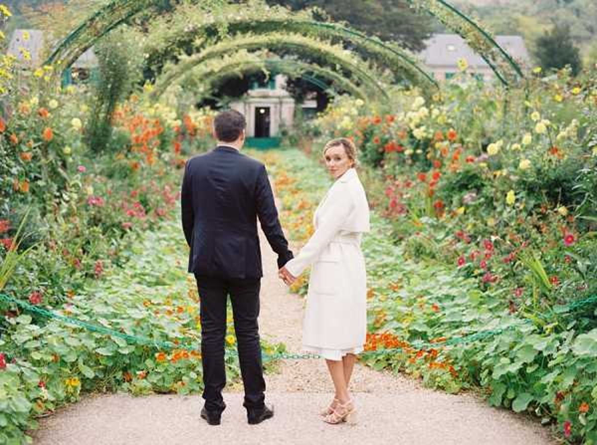 A couple walks hand-in-hand along a gravel garden path lined with dense, colorful flower beds featuring orange, red, yellow, and pink blooms, framed by a series of green metal arched trellises covered in climbing plants. The groom wears a dark navy suit with black shoes, while the bride wears a knee-length white coat dress and nude heeled sandals, glancing back over her shoulder toward the camera. A building with teal-painted woodwork is visible at the far end of the path. The shot is a medium portrait taken from behind the couple, emphasizing the garden archway tunnel composition.