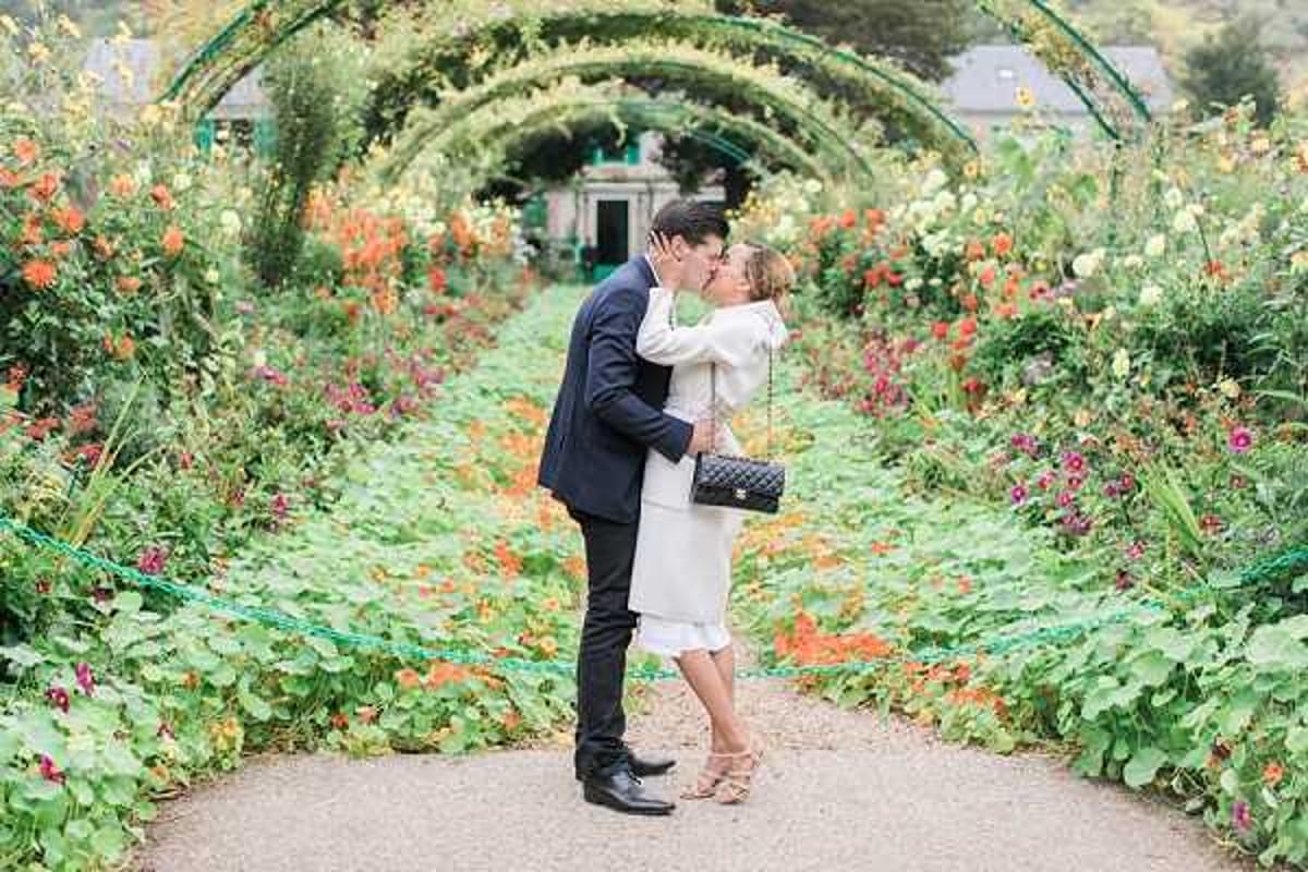 A couple shares a kiss while standing on a gravel path in a lush garden, likely Monet's garden in Giverny, France. The setting features a series of arched trellises covered in climbing plants and flowers in orange, red, yellow, and green tones that create a tunnel-like perspective leading to a small building in the background. The man wears a navy blue suit with dark trousers and black dress shoes, while the woman wears a knee-length white or ivory coat dress with a ruffle hem, nude block-heeled sandals, and carries a quilted black Chanel crossbody bag. This appears to be an engagement or post-wedding portrait session, captured as a full-length couple portrait in natural daylight.