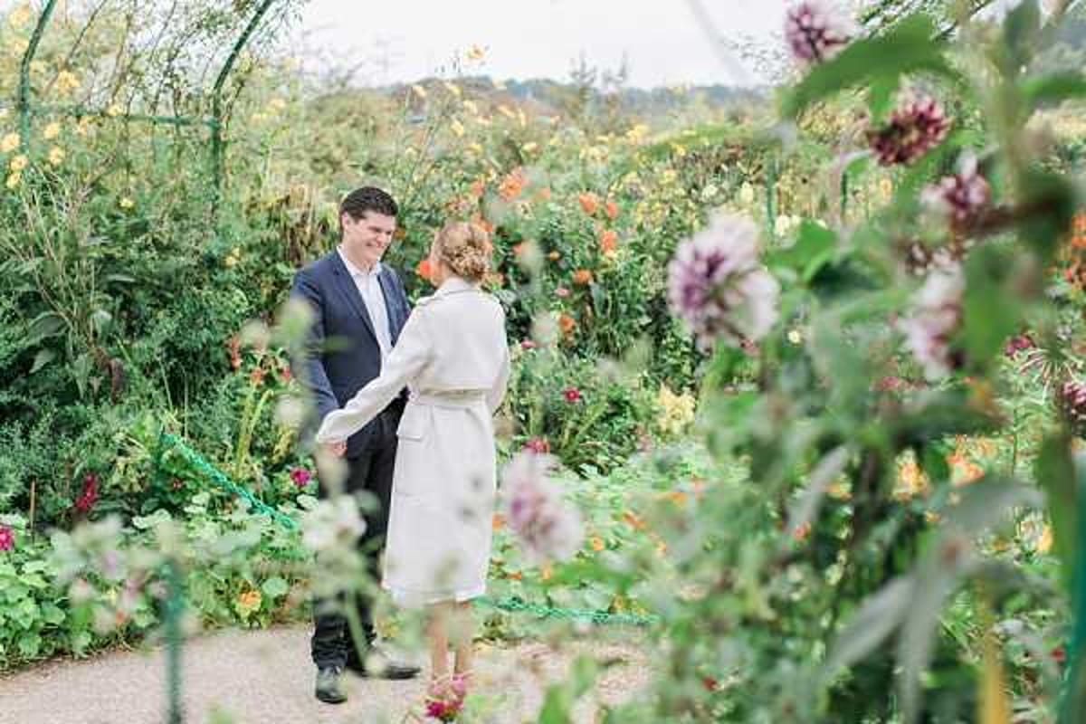 A couple stands facing each other in a lush garden setting, holding hands and smiling. The man wears a dark navy suit with a white shirt, while the woman wears a cream-colored trench coat over what appears to be a light-colored dress, with her hair styled in an updo. The garden is densely planted with tall flowering plants in yellow, orange, and deep red tones, with purple allium blooms framing the foreground. The shot is taken from a medium distance through the garden foliage, creating a layered, framed composition.
