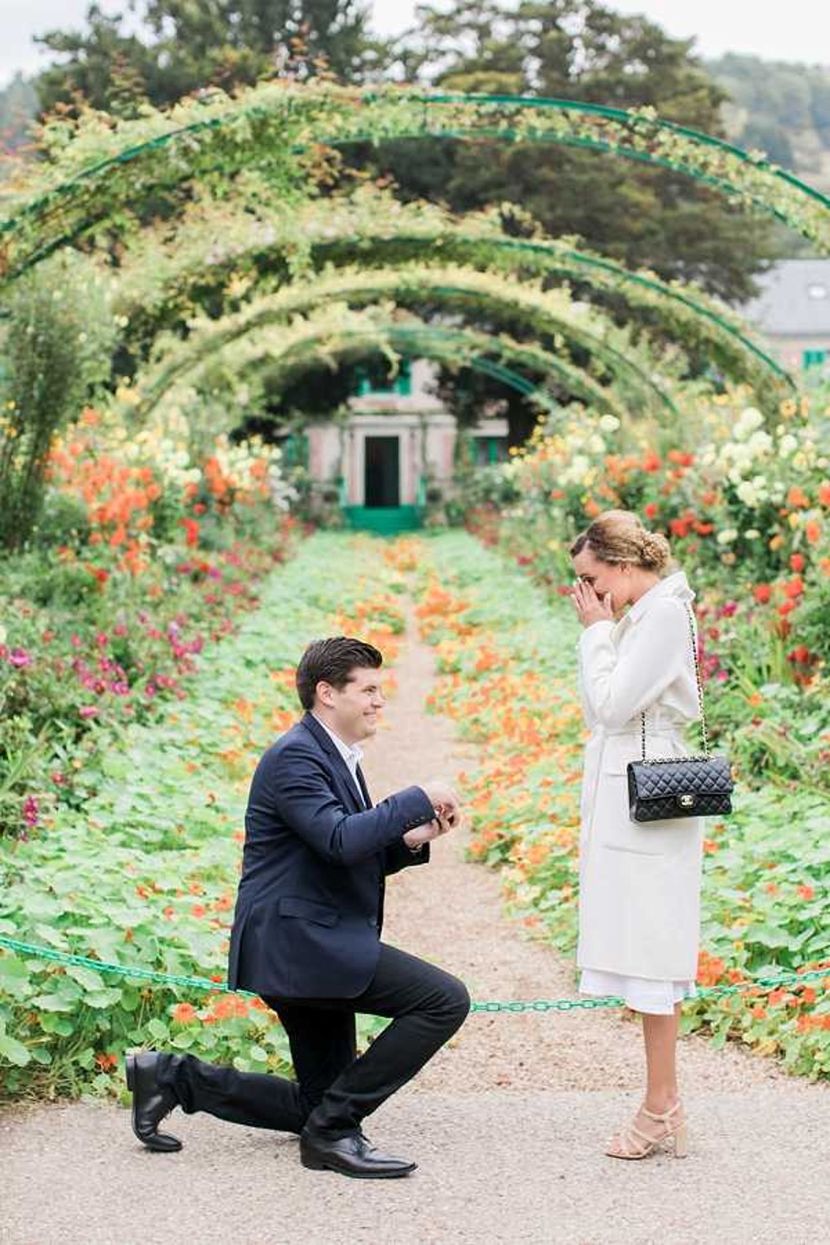 A marriage proposal is taking place in a formal garden, identifiable as Claude Monet's garden in Giverny, with a tunnel of arched trellises covered in climbing plants and blooming orange, red, and yellow flowers lining a gravel path leading toward the iconic green-painted house in the background. A man in a dark navy suit is down on one knee holding a ring box open, smiling up at a woman who is covering her mouth with both hands in a surprised reaction. The woman is wearing a white long coat layered over a white dress, nude strappy heeled sandals, and carrying a black quilted Chanel crossbody bag with gold chain. The shot is a full-length portrait taken from a medium distance, centered on the couple within the flower-lined garden path.