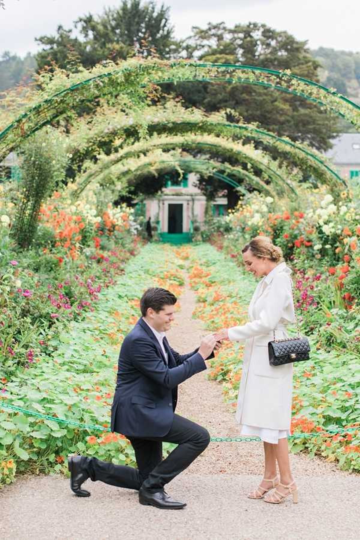 A marriage proposal is taking place outdoors in a formal garden, with a man in a navy suit down on one knee holding the woman's hand as she looks down at him smiling. The woman is dressed in a white coat over a white outfit, wearing nude strappy heeled sandals and carrying a black quilted chain-strap handbag. They are standing on a gravel path lined with arching green metal trellises covered in climbing plants and flowers, with dense garden borders of orange, red, pink, and yellow blooms on either side, and a pink-painted building visible at the end of the path in the background. The composition is a full-length portrait shot taken from a medium distance, capturing both figures in full against the floral archway backdrop.