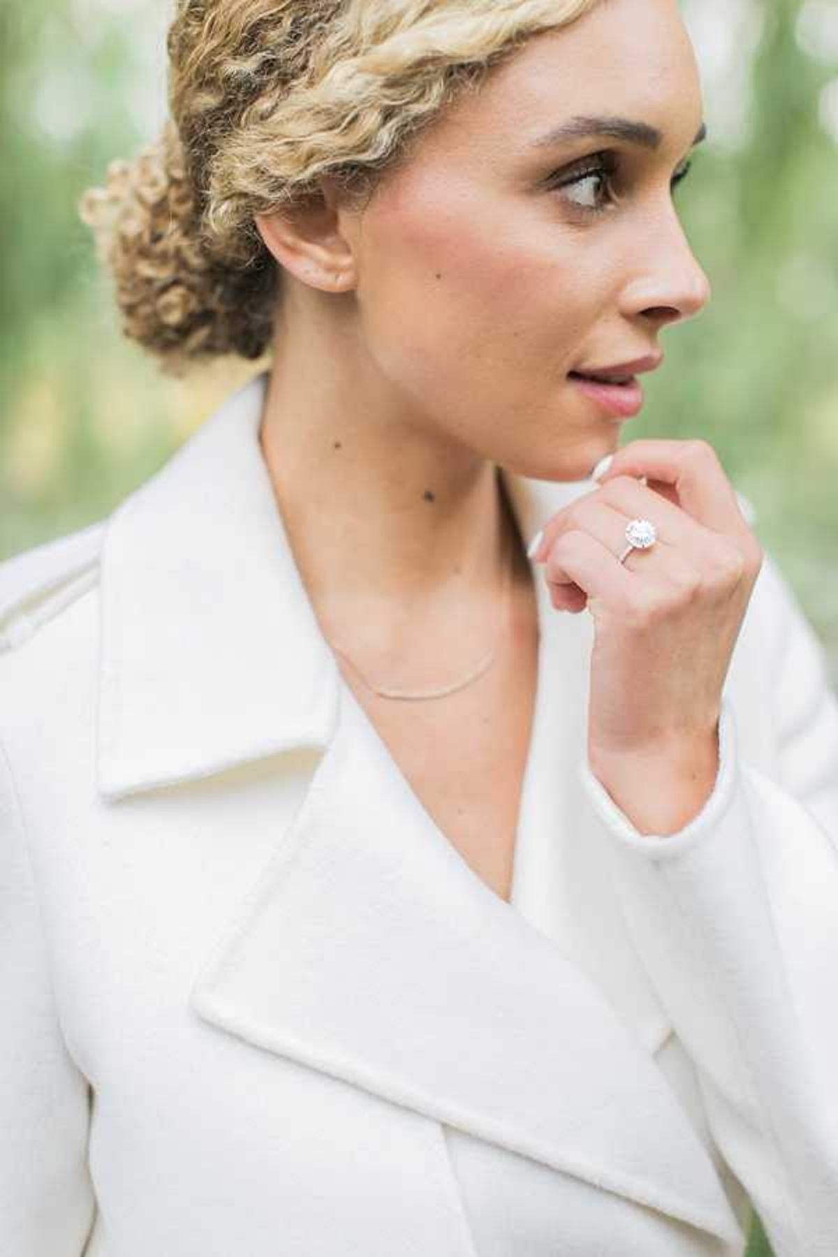 A close-up portrait of a bride with naturally curly blonde hair styled up, wearing an ivory structured coat with wide lapels and a delicate thin silver necklace. Her left hand is raised near her chin, prominently displaying a halo-style diamond engagement ring with a pavé band. She is gazing slightly off-camera with a neutral expression. The background is softly blurred with hints of greenery. The composition is a shallow depth-of-field portrait shot emphasizing the ring and bridal styling details.