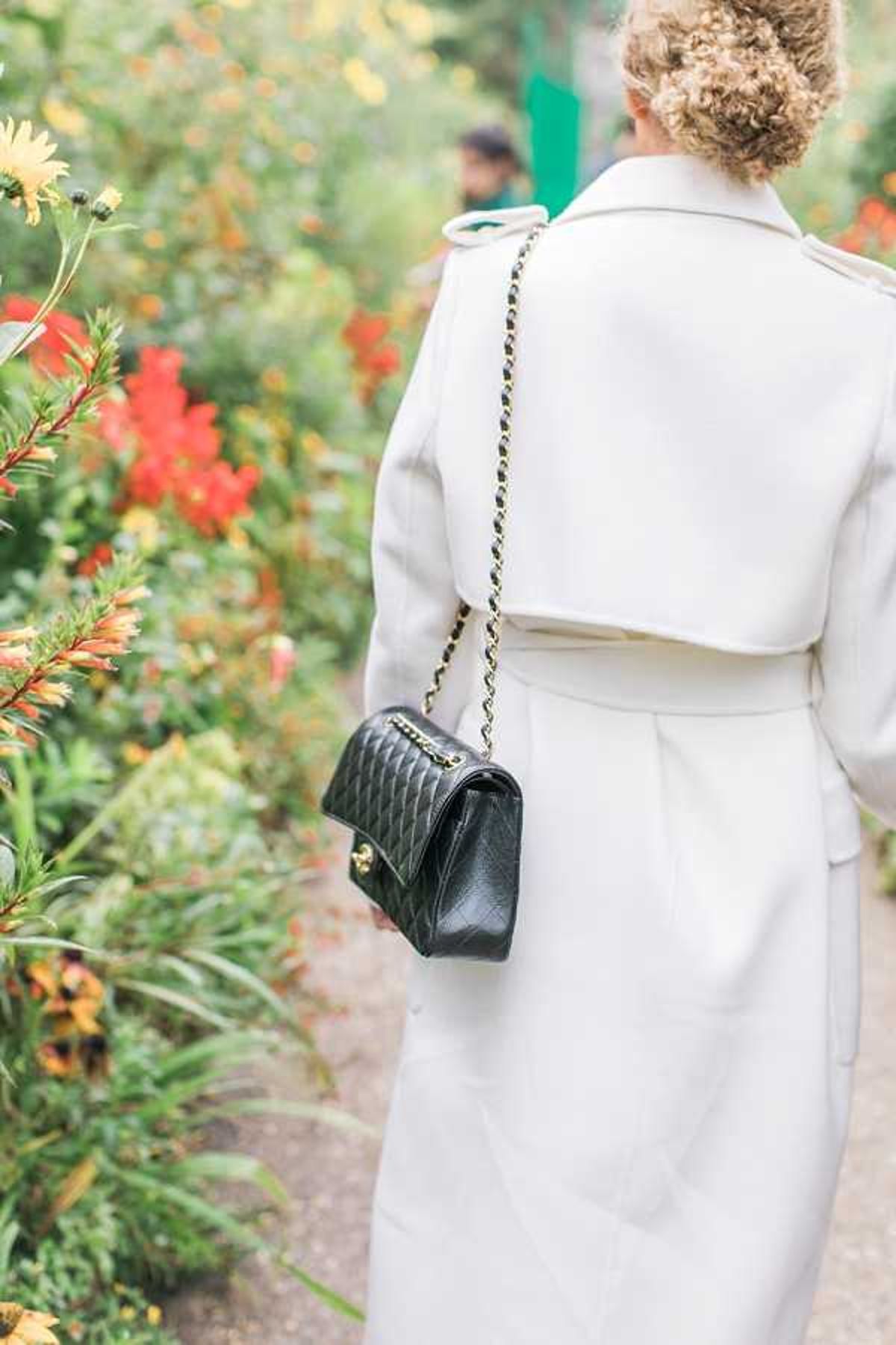 A close-up portrait shot taken from behind of a woman, likely the bride or a wedding guest, wearing a white belted wrap coat with her curly blonde hair styled in an updo. She carries a small black quilted leather handbag with a gold chain strap, consistent with a classic designer style. The setting is an outdoor garden path with colorful red and yellow flowering plants visible in the soft-focus background. The overall styling is classic and polished, with the contrast between the white coat and black bag as a deliberate accessory choice.