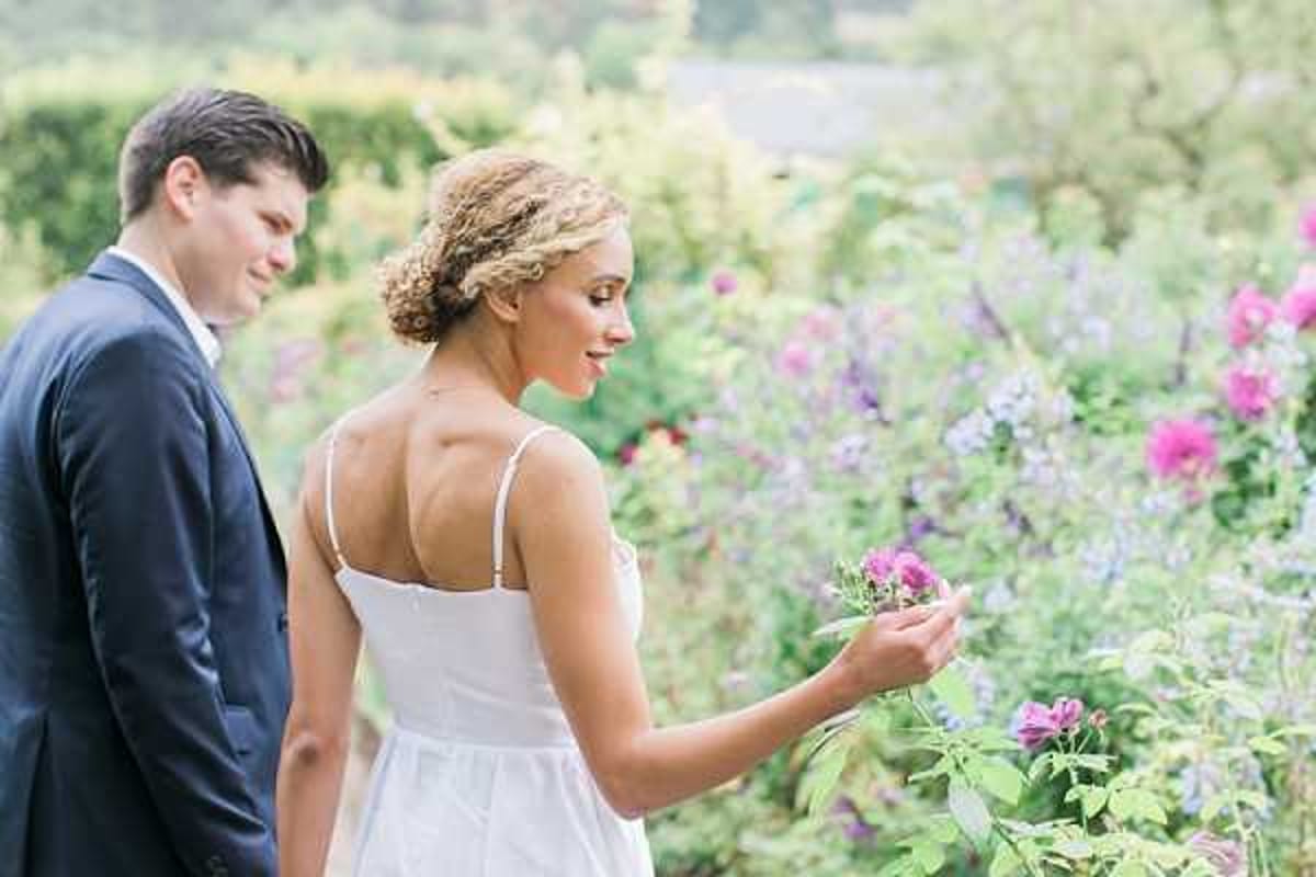 A couple portrait taken outdoors in a flowering garden, with the bride gently touching a deep pink rose on its stem. The bride wears a white spaghetti-strap dress with a low back and has her curly blonde hair pinned up, while the groom stands beside her in a navy suit, looking down toward her. The background is filled with blooming pink roses and lavender-purple flowers, softly out of focus. The shot is a medium close-up taken from behind and slightly to the side, emphasizing the bride's back and the natural garden setting.