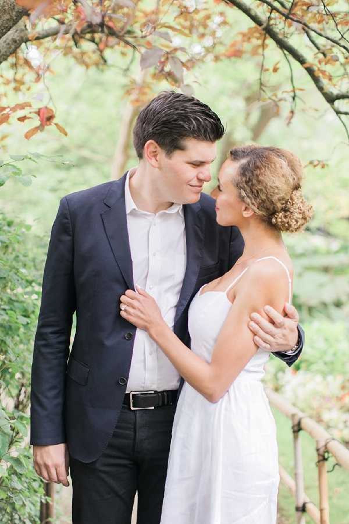 A couple poses together in a garden setting for what appears to be an engagement or pre-wedding portrait session. The man wears a navy blazer over a white dress shirt with dark trousers and a black belt, while the woman wears a simple white spaghetti-strap slip dress with her curly hair styled in an updo. The two face each other closely, with his arm around her shoulders and her hand resting on his lapel. The composition is a mid-length portrait shot with a softly blurred green and rust-toned foliage background.