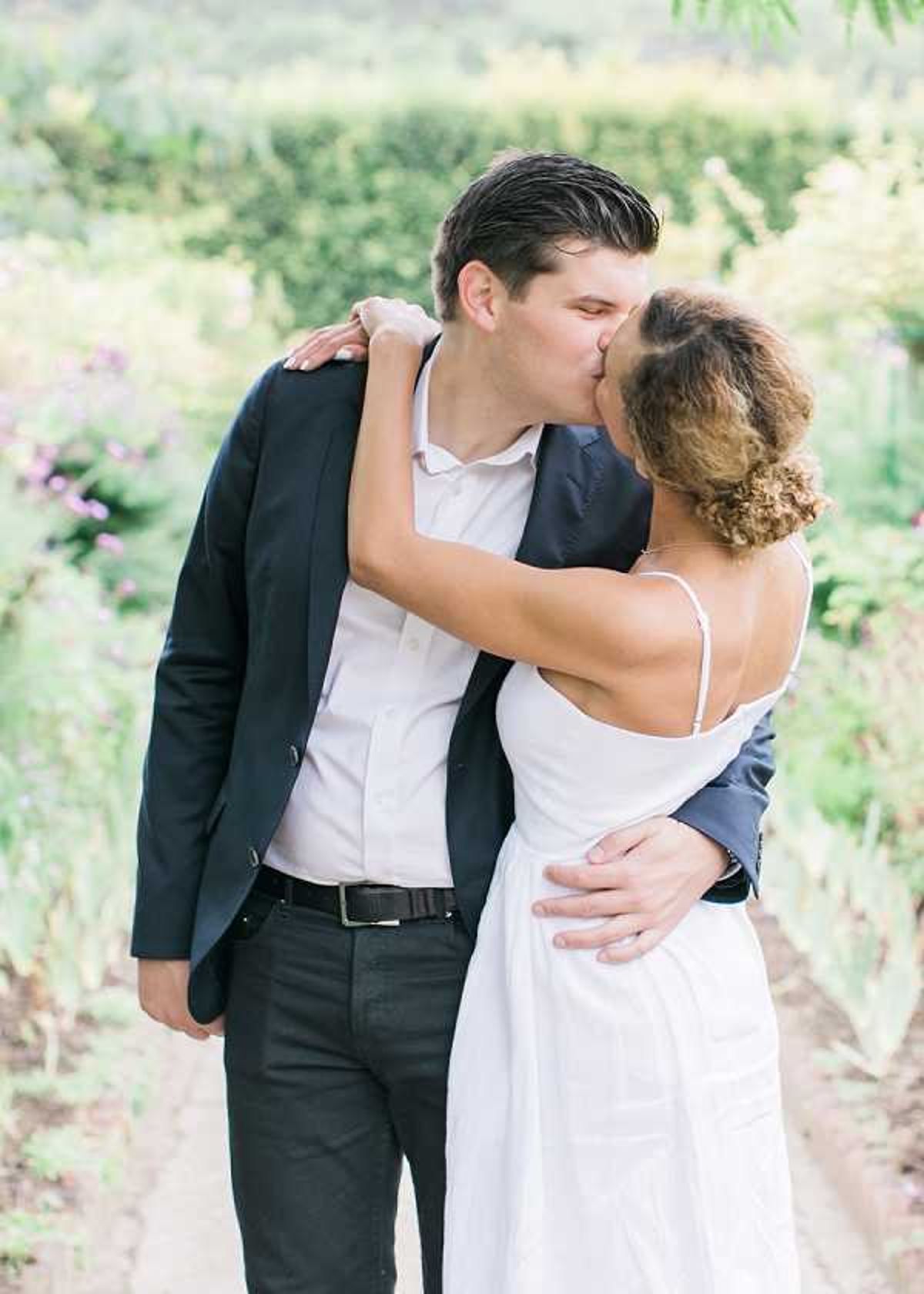 A couple shares a kiss during an outdoor portrait session along a garden path, with lush green hedges and soft purple wildflowers visible in the blurred background. The groom wears a navy blazer, white dress shirt open at the collar, and dark trousers with a black belt, while the bride wears a simple white spaghetti-strap dress with her curly hair worn up. The styling is minimal and casual-modern, with no visible floral or decor elements. The shot is a medium portrait framing both figures from roughly the waist up, with soft natural light and a shallow depth of field.
