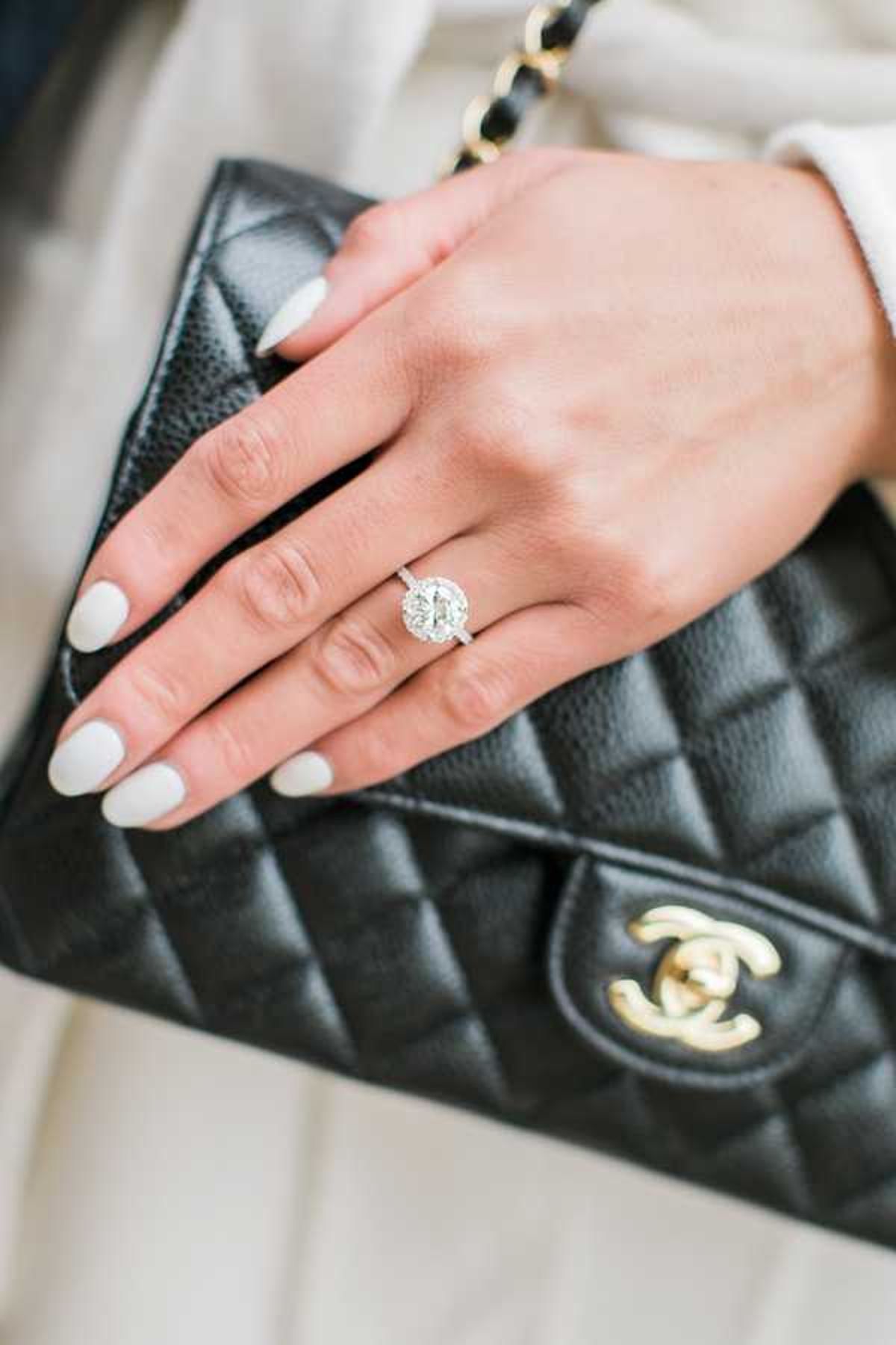 Close-up detail shot of a woman's hand resting on a black quilted leather Chanel handbag with gold hardware. The hand features white-painted nails and a silver halo-style diamond engagement ring with a large round center stone surrounded by a pavé diamond halo and diamond-set band. The woman is wearing a cream or ivory outfit visible in the background. The image is sharply focused on the ring and bag, with a soft-focus background, giving it a polished accessory-detail composition typical of bridal getting-ready coverage.