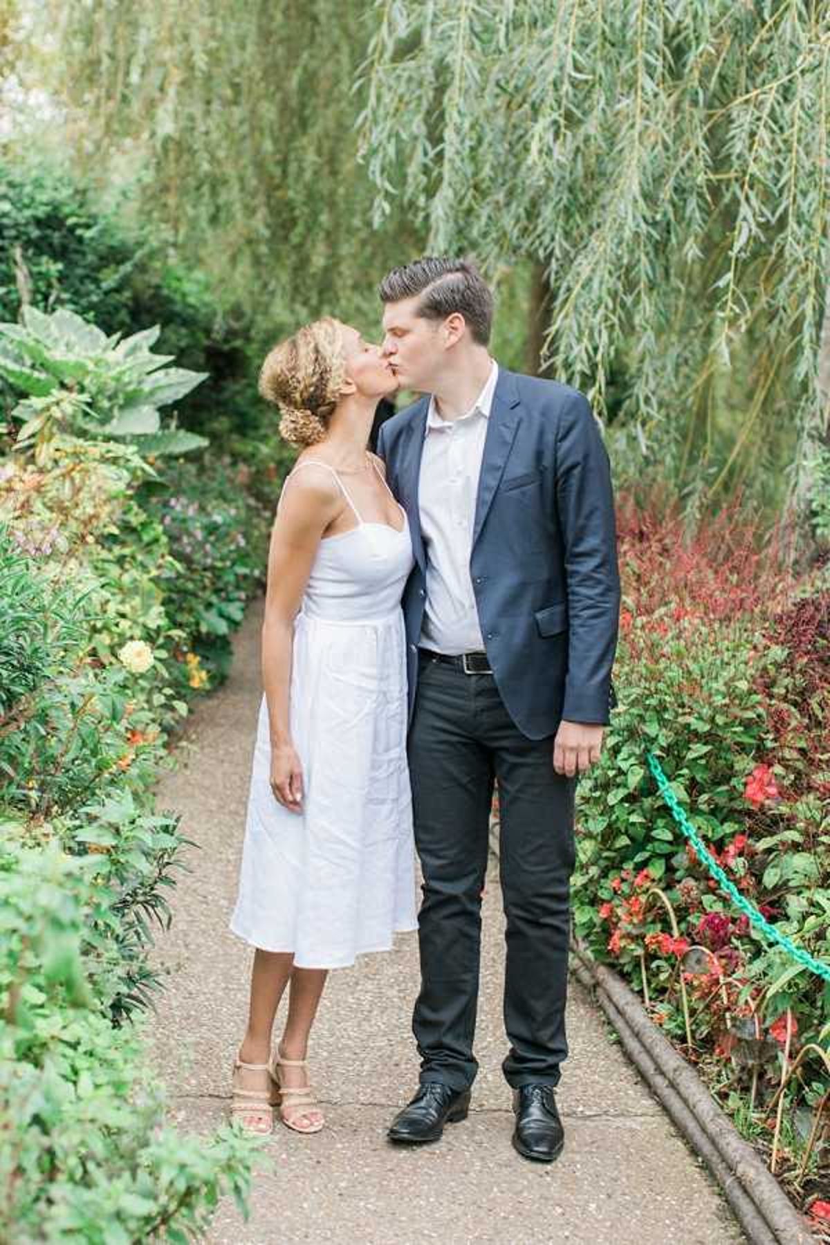 A couple shares a kiss while standing on a garden pathway, photographed in a medium full-length portrait. The bride wears a knee-length white spaghetti-strap dress with a relaxed silhouette and nude strappy heeled sandals, with her hair styled in an updo. The groom wears dark trousers, a white shirt, and a navy blazer with black oxford shoes. The garden setting features lush greenery, red and yellow flowering plants along a bordered path, and trailing willow branches overhead, suggesting a public or estate garden. The overall styling is casual and relaxed rather than formal, with a light, airy color palette.