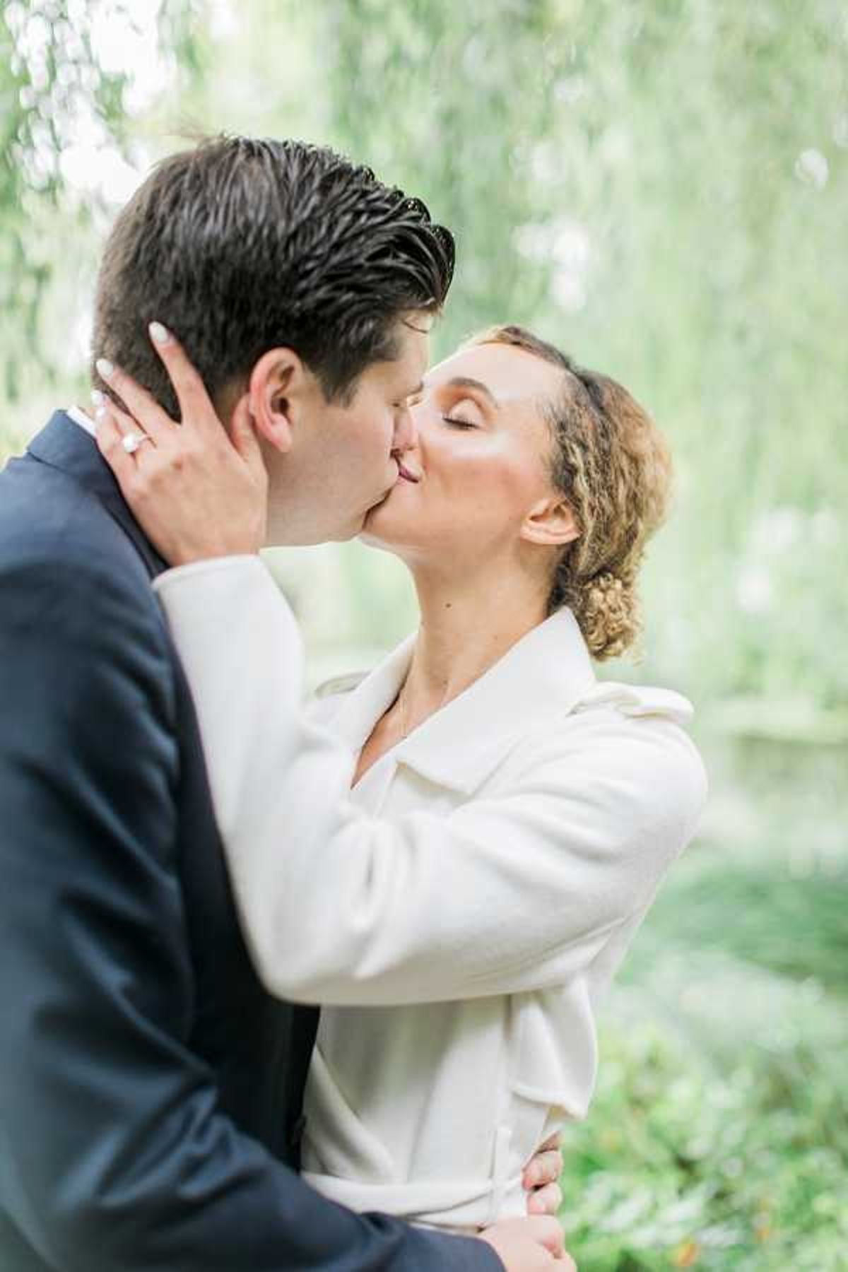 A couple shares a kiss in a close-up portrait shot outdoors. The groom wears a navy suit jacket and the bride wears a white structured coat with a wide lapel collar, with her curly hair styled in an updo. She places one hand on his face, showing a diamond ring. The image is a medium close-up with soft, bright natural light and a blurred green background.