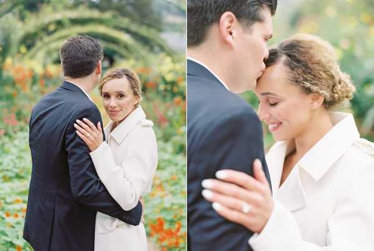 A two-panel couple portrait shot outdoors in a formal garden setting with green arched trellises and orange and yellow flowering plants visible in the background. The woman wears a white structured coat dress and has her hair styled in an updo, while the man wears a dark navy suit; she faces the camera smiling in the left panel while embracing him from behind. The right panel is a closer portrait shot showing the man kissing her on the forehead while she smiles with eyes closed, with her hand — featuring a diamond engagement ring — visible in the foreground. Both shots use a shallow depth of field with the garden softly blurred behind the couple.