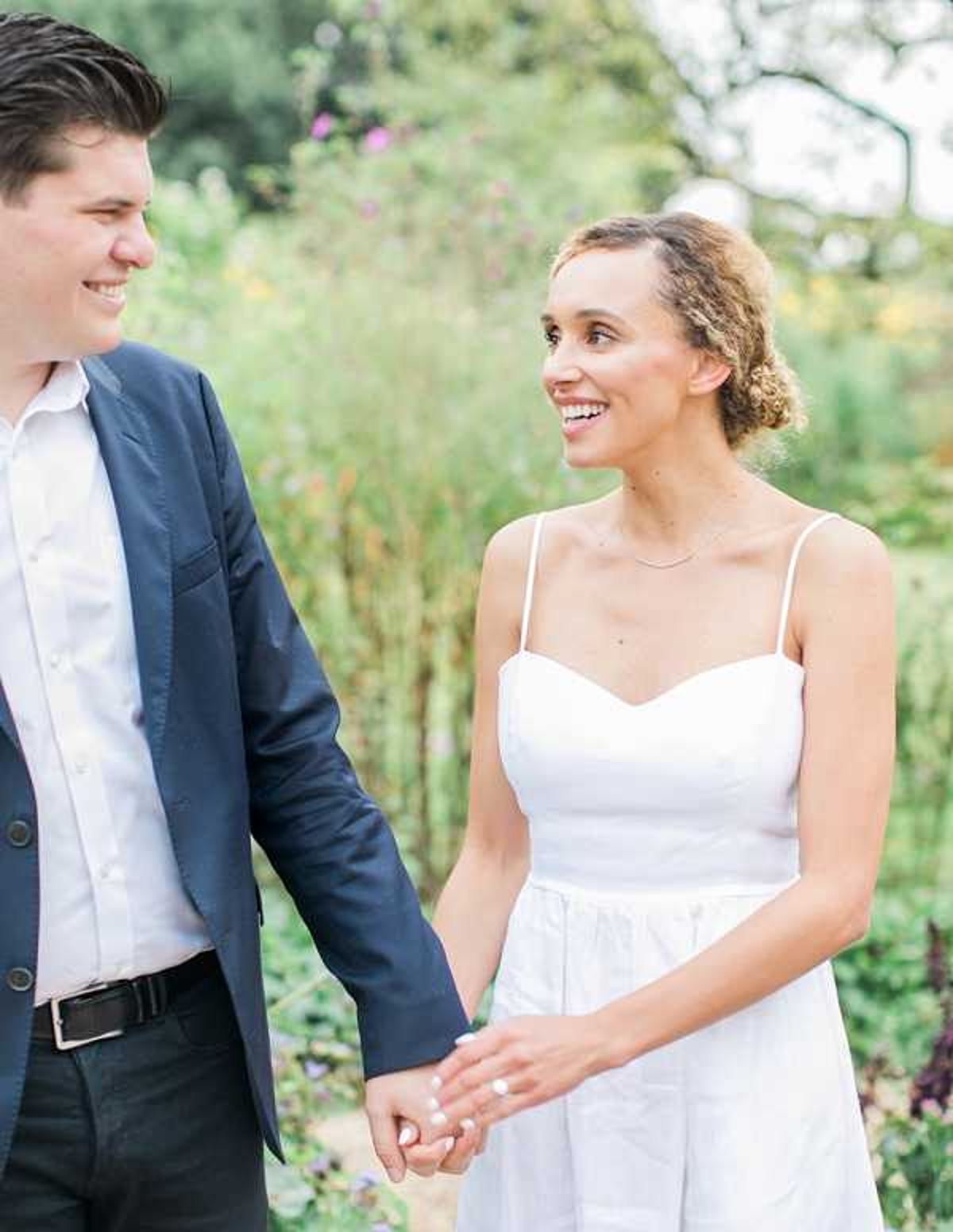 A couple walks hand-in-hand outdoors in a lush garden setting, smiling at each other in a candid portrait. The bride wears a simple white spaghetti-strap dress with a sweetheart neckline and her hair styled in an updo, accessorized with a delicate chain necklace. The groom is dressed in a navy blazer with a white open-collar shirt and dark trousers. The shot is a close-up portrait with a soft, airy feel and a blurred green garden background.