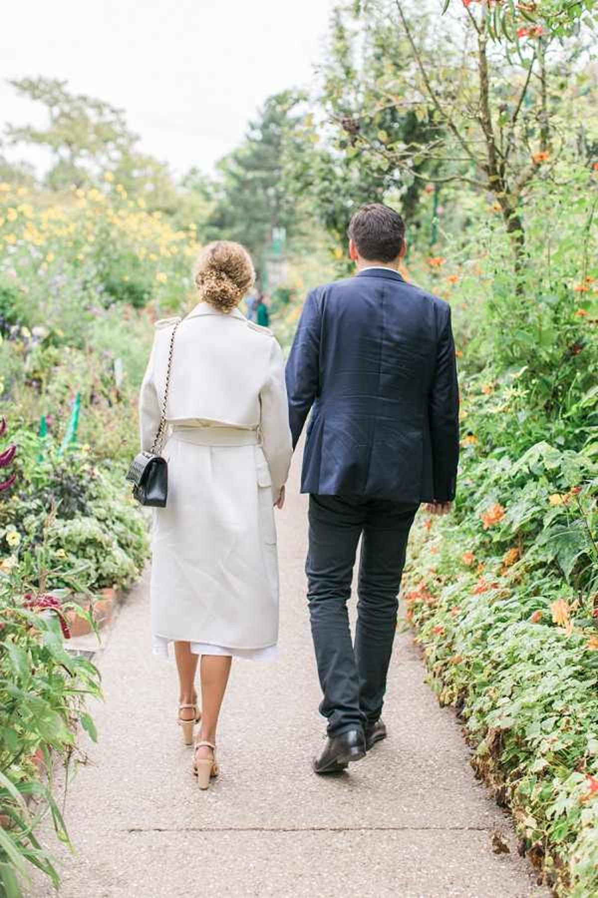 A couple walks hand-in-hand away from the camera along a garden path lined with dense, colorful plantings. The woman wears a cream belted coat over a white dress, nude block-heel sandals, and carries a small black quilted crossbody bag, with her hair styled in an updo. The man wears a navy suit jacket with dark trousers and black shoes. The setting is an outdoor formal garden with a wide gravel or paved pathway bordered by abundant flowering plants. The shot is a full-length portrait taken from behind, with soft natural light and a slightly overcast sky visible above the treeline.
