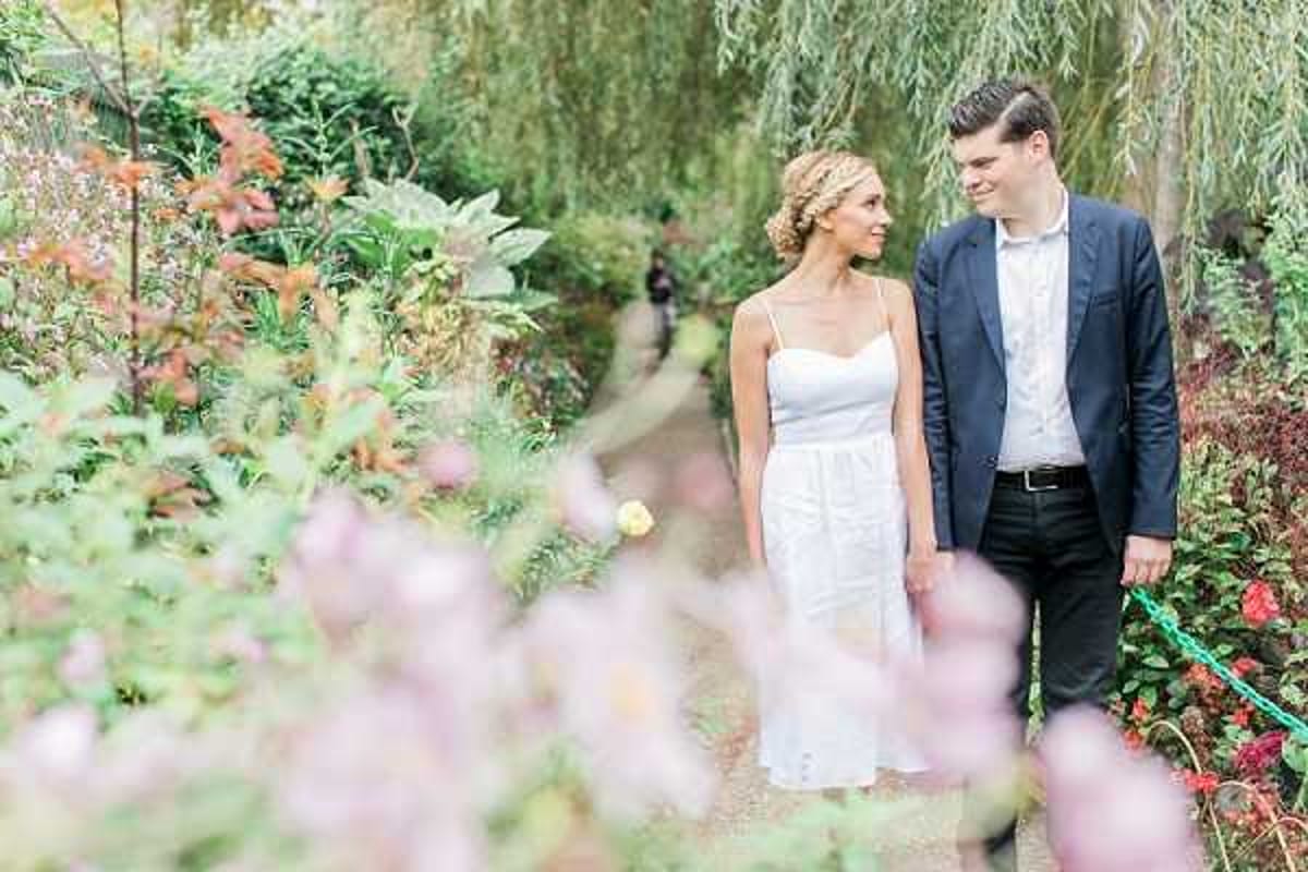 A couple walks together along a garden path, with the bride looking toward the groom and both sharing a smile. The bride wears a simple white spaghetti-strap midi dress with a sweetheart neckline and has her hair styled in a braided updo, while the groom wears a navy blazer with dark trousers and a light blue shirt. The outdoor setting is a densely planted garden path lined with varied foliage in green, burgundy, and amber tones, with soft pink and red flowering plants in the foreground creating a natural frame. The shot is a medium portrait with a shallow depth of field, giving the blurred foreground blooms a soft, painterly quality, and the overall image has a bright, airy, light-and-airy editing style.