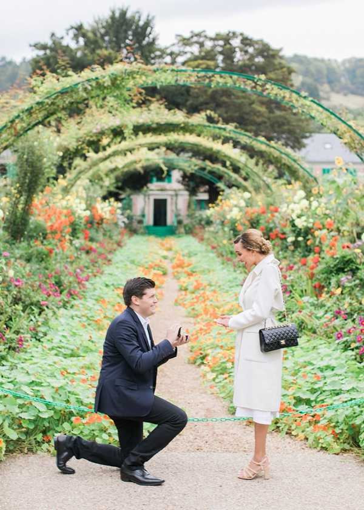 A marriage proposal is taking place outdoors in a formal garden, identifiable as Monet's garden in Giverny, France. A man dressed in a navy suit is down on one knee holding open a ring box toward a woman wearing a white coat, a white midi skirt, strappy heeled sandals, and carrying a black quilted Chanel crossbody bag. The garden's iconic arched metal tunnels are covered in foliage and flowering plants in orange, red, yellow, pink, and white, creating a tunnel perspective leading to a green building in the background. The shot is a full-length portrait framing both subjects in the foreground with the flower-lined gravel path and arched tunnel as the backdrop.