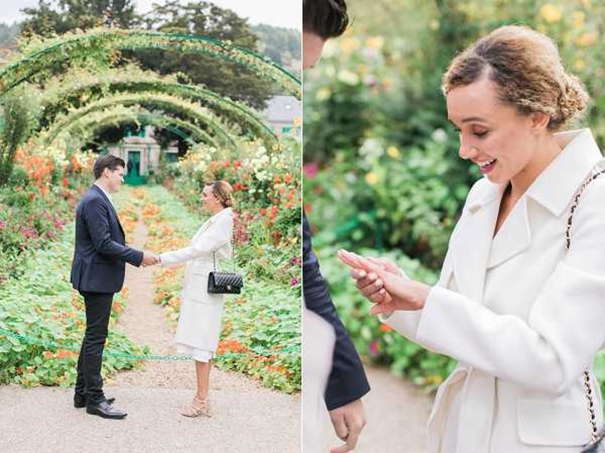 A two-panel engagement or proposal image taken outdoors in a formal flower garden with arched green metal trellises covered in climbing plants forming a tunnel-like pathway, bordered by dense plantings of orange, red, and green flowers. In the left panel, a wide shot shows a man in a navy suit holding hands with a woman in a white knee-length coat and nude heeled sandals, carrying a black quilted chain-strap handbag; they face each other on a gravel path. In the right panel, a close-up portrait captures the woman's joyful expression as she looks down at her hand — likely just after receiving a ring — wearing the same white coat, her hair styled in an updo with a braided detail.