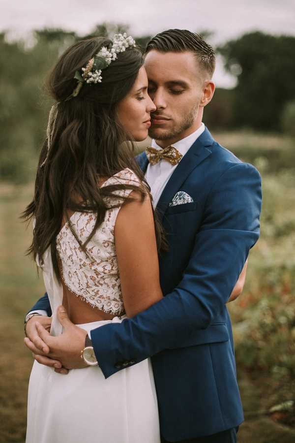 A close-up portrait of a couple embracing outdoors in a natural, open field setting with soft bokeh background. The bride wears a backless lace gown with a low-cut open back and has long dark hair styled half-up, adorned with a floral crown of small white blooms and greenery. The groom wears a cobalt blue suit jacket with a white dress shirt, a gold patterned bow tie, and a blue floral pocket square. The couple's eyes are closed and their faces are close together in an intimate pose. The overall styling has a boho aesthetic. Portrait composition with a shallow depth of field.