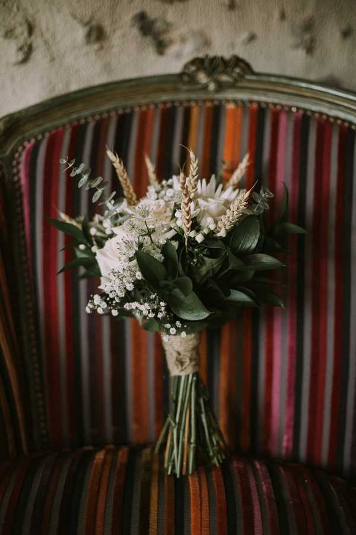 Close-up detail shot of a bridal bouquet propped upright on the seat of a vintage French armchair with a multicolored striped fabric in red, orange, burgundy, and dark tones, accented with brass nail-head trim and an ornate gilded frame. The bouquet is composed of white roses, baby's breath, deep green eucalyptus and mixed foliage, and dried golden wheat stalks, giving it a rustic, organic style. The stems are wrapped in what appears to be ivory lace or ribbon. The background wall has a textured, aged plaster finish, reinforcing the rustic French interior setting.