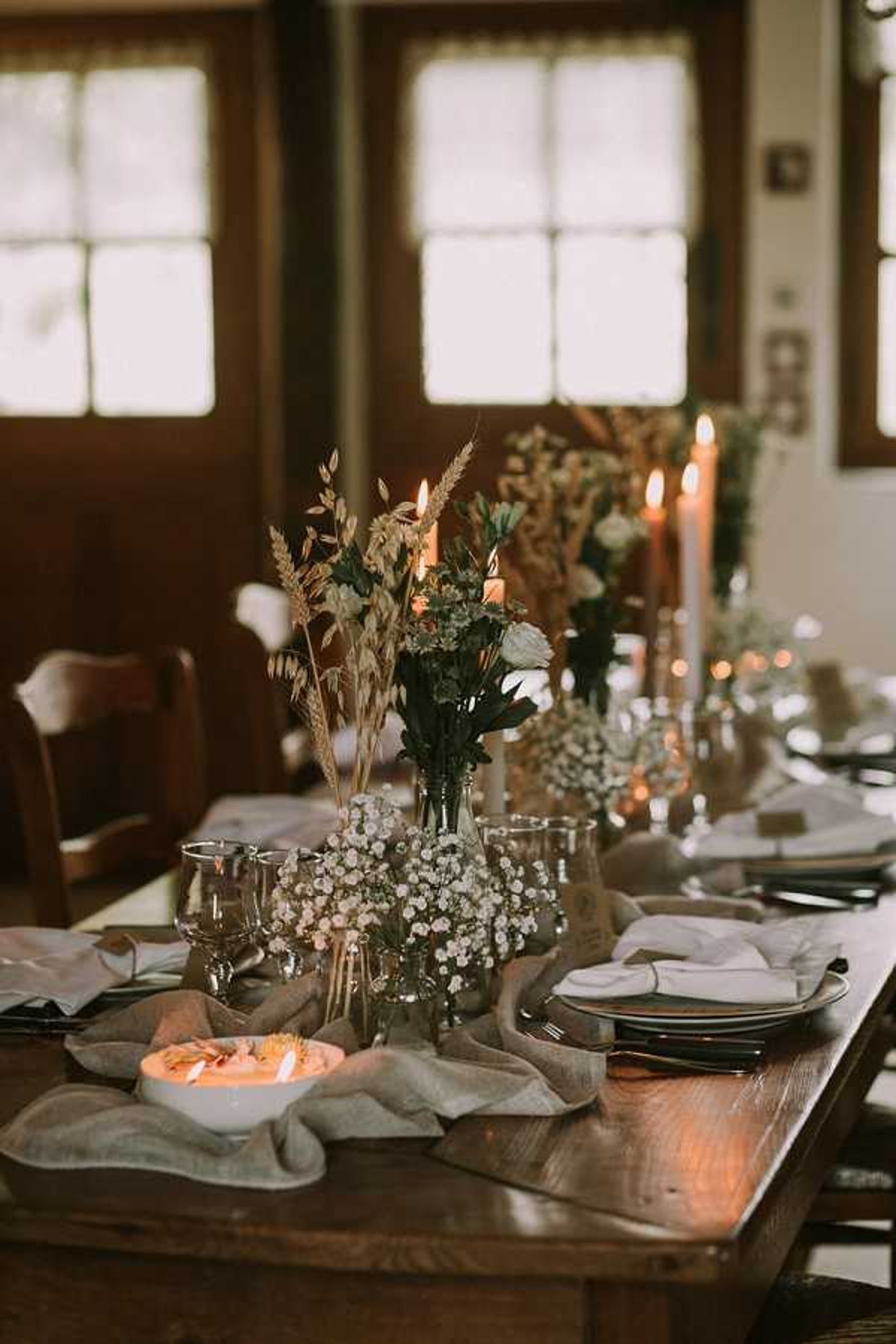 A close-up detail shot of a rustic wedding reception tablescape set on a dark wood farm table in an indoor venue with wood-framed windows visible in the background. The centerpiece arrangement features clusters of white baby's breath, dried wheat stalks, greenery, and a single white rose displayed in glass bud vases of varying heights, styled in a relaxed, boho-rustic aesthetic. Tall terracotta-toned taper candles are lit along the table runner, which is made of loosely draped linen in a muted grey-green tone. Place settings include dark charger plates layered with white linen napkins, wine glasses, and black flatware, with a small white bowl containing an orange-toned appetizer visible in the foreground.