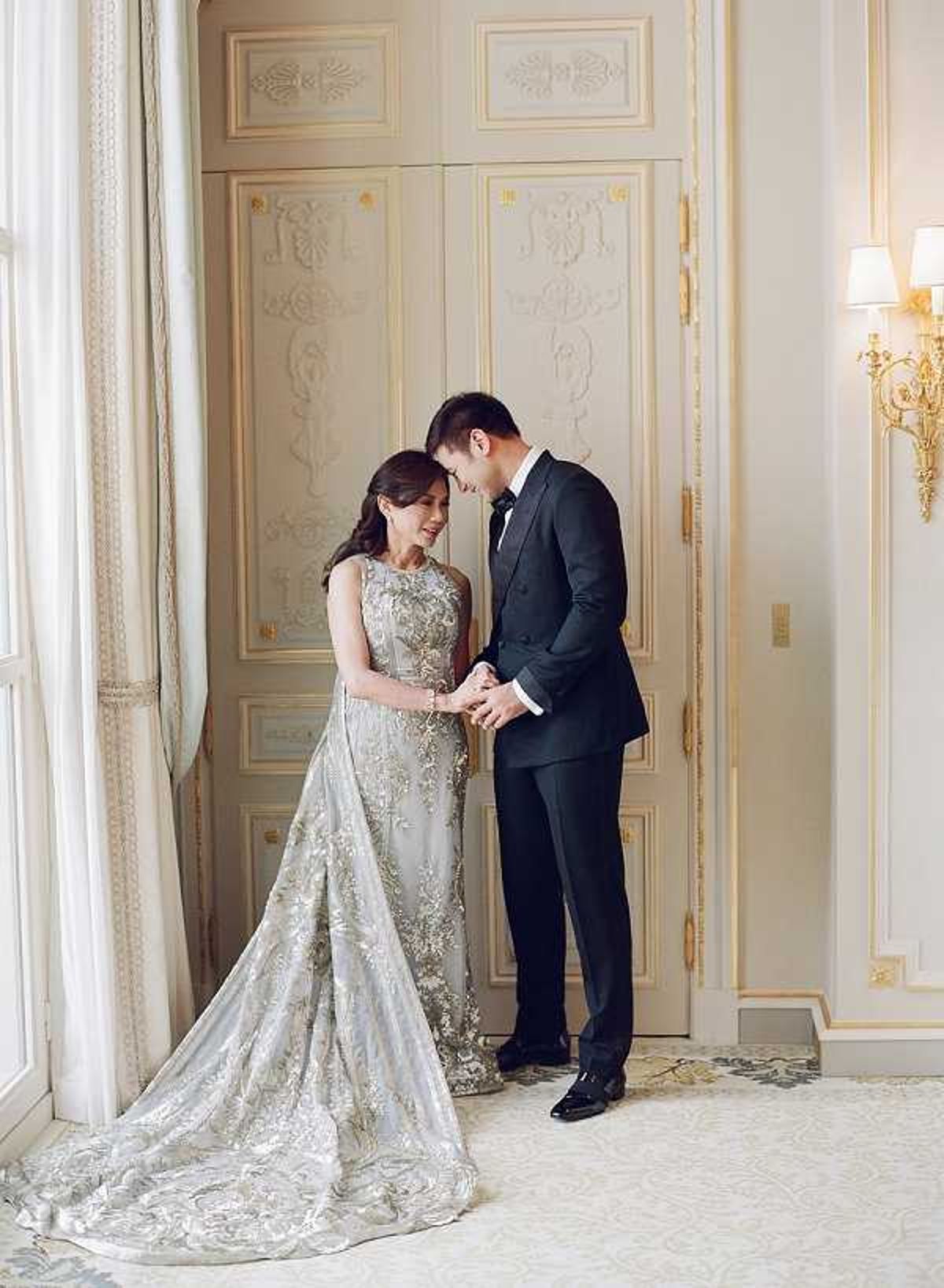 A couple shares an intimate portrait moment indoors, standing close together with foreheads nearly touching and hands held. The bride wears a silver-grey heavily embroidered gown with gold floral detailing, a fitted bodice, and a full train spread across the floor; the groom wears a black tuxedo with a black bow tie. They are positioned in front of ornate cream-and-gold double doors with carved decorative panels, characteristic of French classical interior architecture, with a gold wall sconce visible to the right and sheer white drapes framing a tall window to the left. The decor palette is cream, ivory, and gold with a patterned carpet underfoot, reflecting a formal classic French interior style. Medium full-length portrait shot.