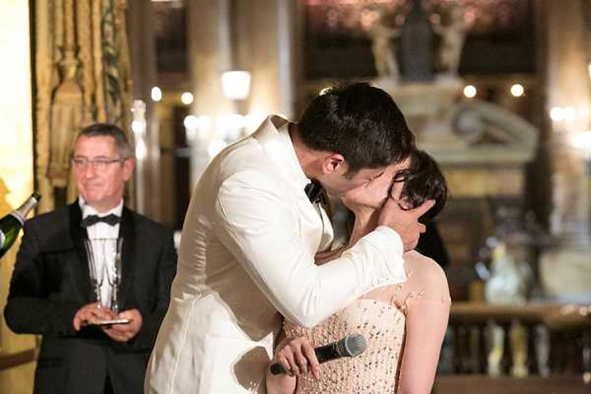 The groom, wearing a white dinner jacket with a black bow tie, leans in to kiss the bride during what appears to be a reception speech moment, as he holds a microphone in one hand. The bride is wearing a blush/champagne sequined or beaded strapless gown. In the background, a man in a black tuxedo and bow tie watches the moment, holding a champagne flute and bottle, likely a wedding host or MC. The setting is an ornate indoor reception hall with warm ambient lighting, gold drapery, and an elaborate decorative fountain or sculptural piece visible in the background, suggesting a classic French chateau or palace-style venue. The shot is a medium close-up portrait capturing the candid kiss.