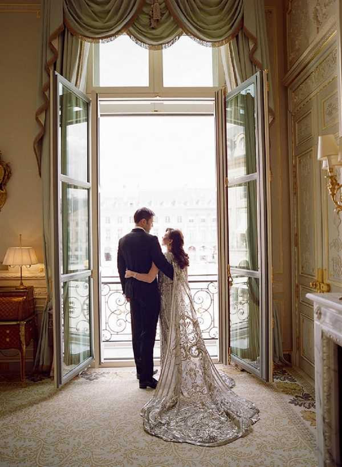 A couple portrait taken indoors in a grand Parisian hotel suite, with the bride and groom standing together facing open French doors that lead to a wrought-iron balcony overlooking a Haussmann-style cityscape. The groom wears a classic black tuxedo, while the bride wears a heavily embroidered silver and ivory long-sleeved gown with an extended cathedral train featuring intricate metallic scrollwork detail. The room features sage green draped curtains with swag valances, gold-accented moldings, a white marble fireplace, wall sconces, and a patterned cream carpet, reflecting a formal classic Parisian interior style. The image is a full-length wide shot, backlit by natural light flooding through the open doors, creating a silhouetted effect against the bright city view beyond. Potential venue feature image.