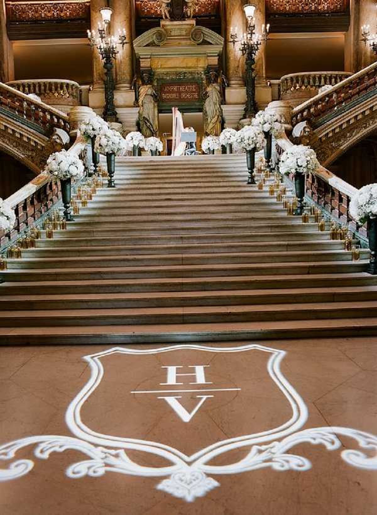 A wide-angle interior shot of the grand staircase at the Palais Garnier (Paris Opera House), decorated for a wedding reception or ceremony entrance. The marble staircase is flanked on both sides by tall dark pedestal urns filled with full white floral arrangements, likely white hydrangeas and roses, interspersed with small gold votive candle holders lining the balustrades. A monogram gobo light projection on the marble floor in the foreground displays the initials 'H' and 'V' inside an ornate shield-shaped crest design. The venue's gilded bronze candelabras, classical sculpture figures, and warm amber lighting from wall sconces illuminate the grand Beaux-Arts architecture. The decor palette is white, gold, and dark green, with a classic, formal styling theme. Potential venue feature image.