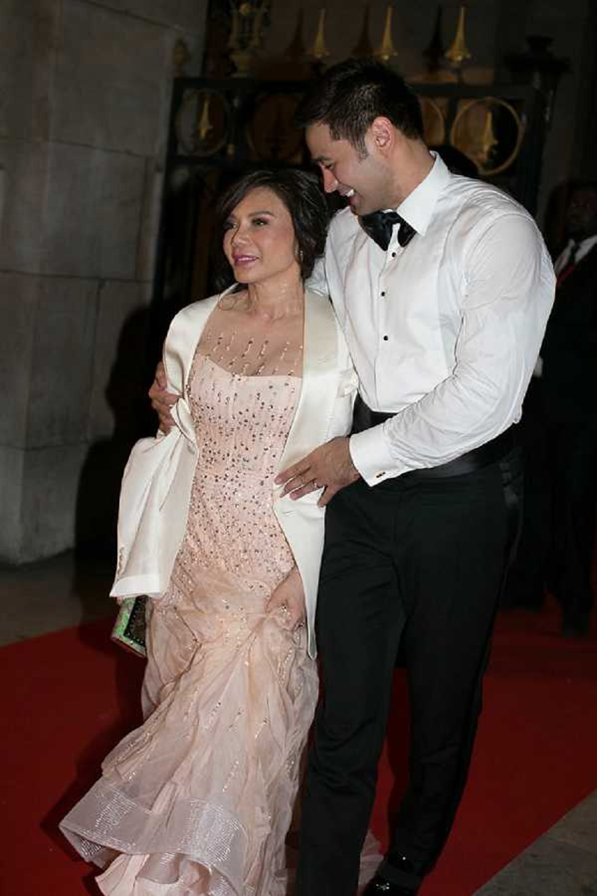 Two wedding guests arrive at what appears to be a formal indoor venue, walking on a red carpet at the entrance. The woman wears a blush pink beaded gown with a mermaid silhouette and a white satin wrap jacket, carrying a small embellished clutch, while the man wears a white dress shirt with a black bow tie and black trousers in a classic tuxedo style. The man has his arm around the woman and they are both smiling. The shot is a medium portrait taken at the venue entrance, with ornate dark architectural details visible in the background.