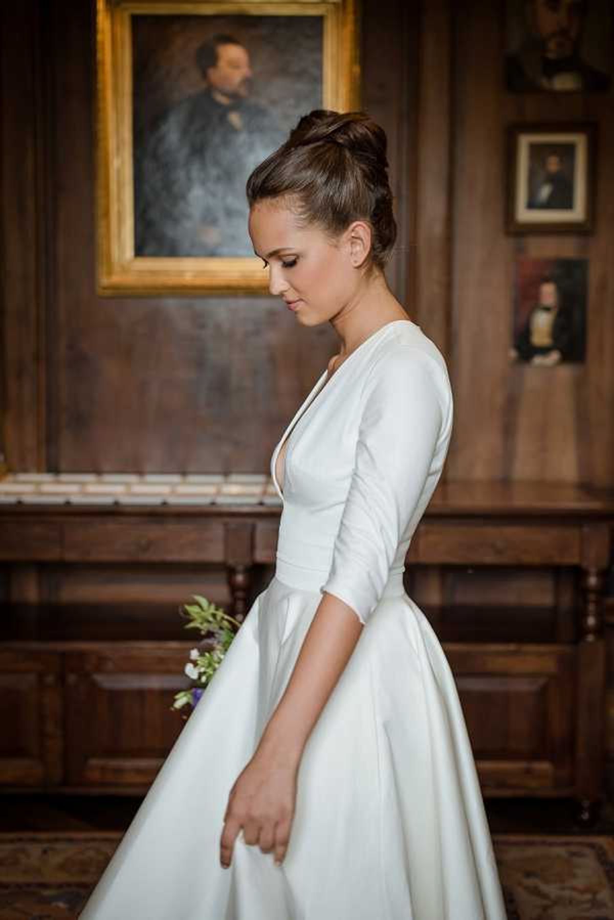 A bridal portrait taken indoors in what appears to be a wood-paneled room of a historic chateau or manor house, with oil portrait paintings in gilt frames visible on the dark wooden walls in the background. The bride stands in profile, looking downward, wearing a white satin ball gown with a deep V-neckline and three-quarter-length sleeves, styled in a clean, modern silhouette. Her dark hair is worn in a high, sleek upknot, and she holds a small loose bouquet featuring purple and green flowers partially visible at her side. The composition is a medium portrait shot with warm, low ambient lighting emphasizing the rich wood tones of the interior.