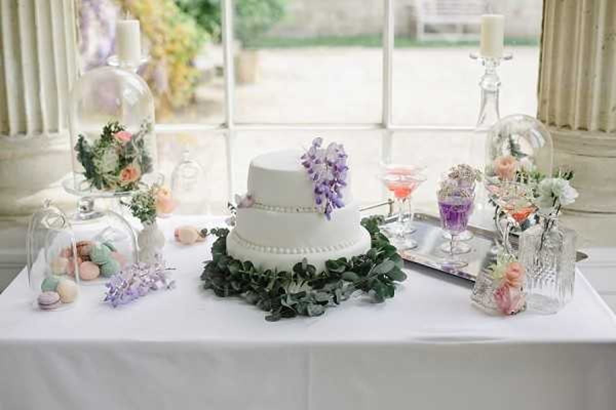 A detail shot of a wedding dessert table set up indoors, likely inside a chateau or manor house with large Georgian-style windows visible in the background. The centerpiece is a two-tier white fondant cake decorated with pearl-dot trim, a wreath of eucalyptus leaves at its base, and a cascade of purple wisteria sugar flowers on top. Surrounding the cake are glass cloches displaying pastel macarons in mint, pink, and lavender, as well as floral arrangements featuring peach and blush roses. A silver tray holds crystal and colored glassware filled with pink and purple drinks or desserts, and additional floral stems in a crystal vase are placed to the right. The overall decor palette is soft pastel — lavender, blush, mint, and white — suggesting a romantic, garden-inspired styling theme. The table is covered with a plain white linen cloth.