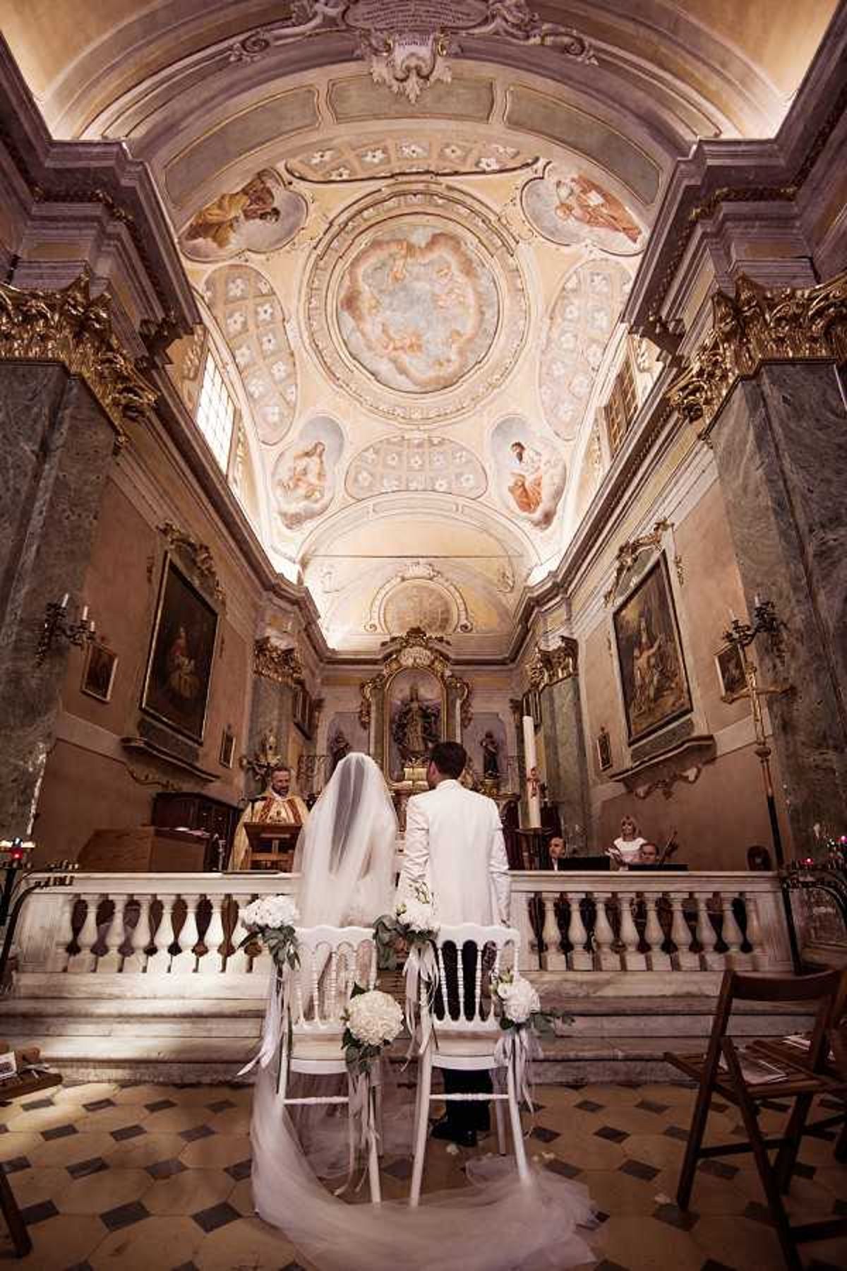 A wedding ceremony taking place inside a historic Baroque chapel with an ornate frescoed ceiling featuring classical figurative paintings and gilded architectural detailing. The bride and groom are seated with their backs to the camera at white chairs decorated with white hydrangea and eucalyptus floral arrangements tied with white ribbon, facing the altar where an officiant in gold vestments stands. The bride wears a long cathedral-length veil and a white gown, while the groom wears an all-white suit. A wide-angle shot captures the full interior from the nave to the altar, emphasizing the vaulted painted ceiling, marble balustrade, candle sconces, and large religious paintings on the side walls. The ceremony style is classic and formal, set within an ornate religious interior with warm ambient lighting. Potential venue feature image.