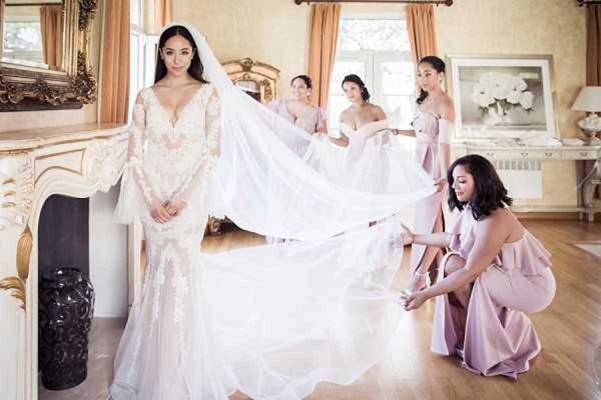 A getting-ready scene photographed indoors in an ornate room featuring a white carved fireplace, gold-framed mirror, and warm peach-toned curtains with hardwood flooring. The bride stands to the left wearing a long-sleeve, deep V-neck fitted lace gown with floral appliqué detailing and a long cathedral veil, which four bridesmaids are arranging and lifting behind her. The bridesmaids are dressed in dusty rose/mauve off-shoulder and ruffle-back satin gowns. The composition is a wide portrait-style shot capturing both the bride in the foreground and the bridesmaids working with the veil in the background, creating depth across the frame.