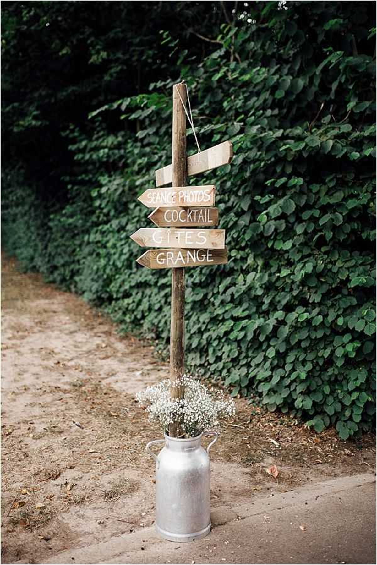 A rustic wedding directional signpost stands along a sandy path at an outdoor venue. The wooden post holds four hand-painted arrow signs in white lettering pointing guests toward 'Séance Photos,' 'Cocktail,' 'Gîtes,' and 'Grange.' At the base of the post, a silver milk churn serves as a vase holding a bouquet of white gypsophila (baby's breath). The overall styling is rustic-country, and the detail shot is taken at medium distance with a shallow depth of field.