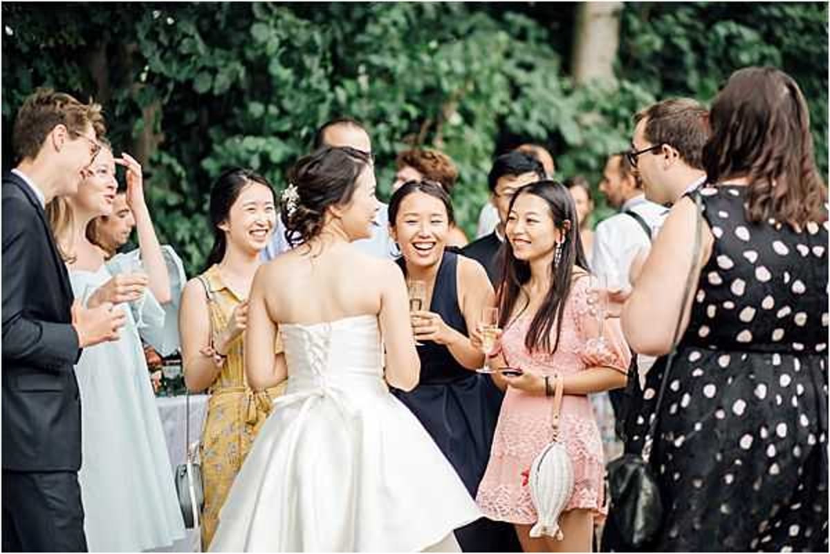 An outdoor cocktail hour scene showing a bride in a strapless ivory ballgown with a lace-up back chatting and laughing with a group of approximately ten guests. The setting appears to be a garden or park with dense green foliage in the background. Guests are dressed in a mix of outfits including a navy dress, a pink lace romper, a yellow floral dress, a mint dress, and a black polka-dot dress, several holding champagne flutes. The mood is relaxed and celebratory, with the bride facing away from the camera toward her guests. Medium wide shot capturing candid interaction among the group.