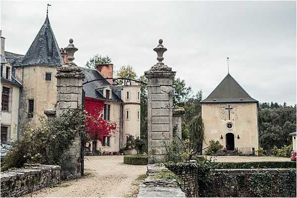 Wide exterior shot of a French chateau complex featuring a grand stone entrance gate with two decorative pillar finials leading to a gravel courtyard. The property includes a multi-story chateau with a pointed slate turret on the left, a cylindrical tower, and a small chapel or dovecote structure with a cross on the right side. No people are visible in the frame. The architecture is classic French rural chateau style with pale stone facades and dark slate roofing. The image has a muted, overcast color palette. Potential venue feature image.