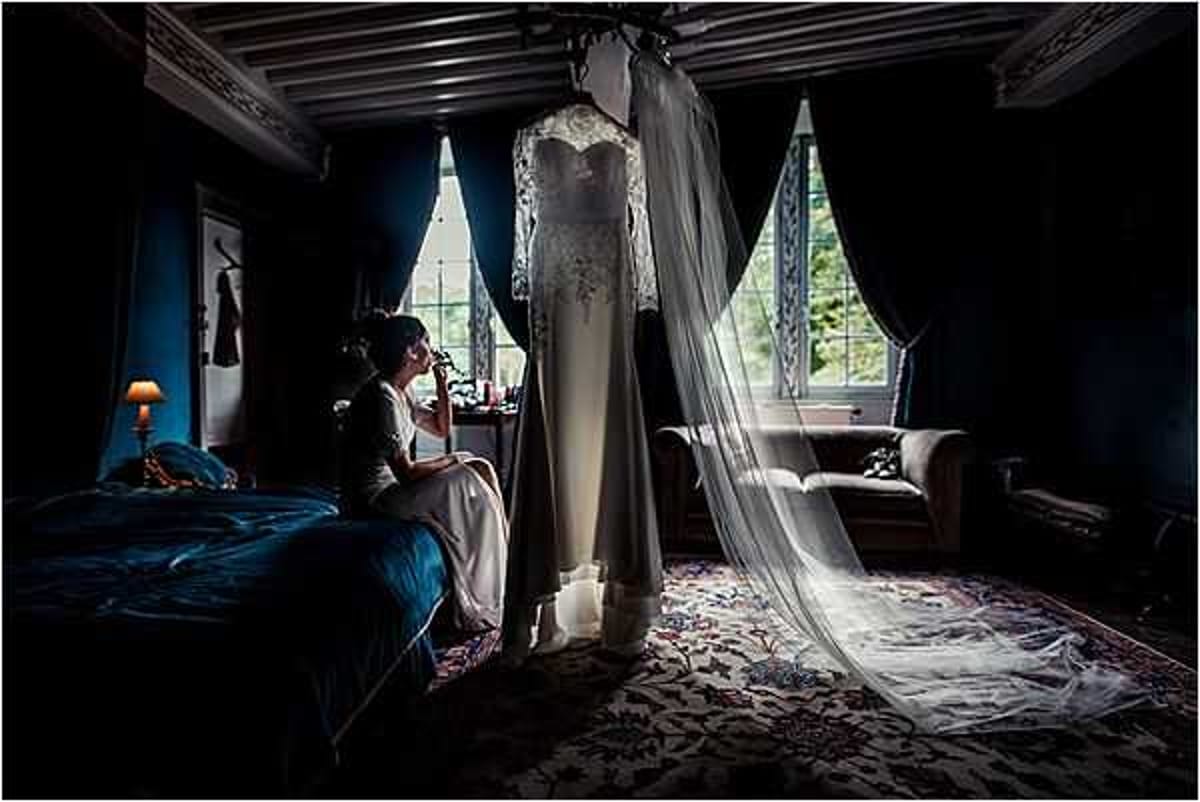 A bride sits on the edge of a bed in a darkly decorated bedroom, appearing to apply makeup or touch her face while looking toward a window. The room features deep teal-blue walls, dark wood ceiling beams, and a richly patterned area rug, suggesting a historic chateau interior. An ivory lace wedding gown with a long flowing veil is hung from the ceiling in the center of the frame, dramatically backlit by natural light streaming through tall curtained windows. The wide-angle shot uses strong contrast between the dark room and the bright windows, creating a moody, dramatic composition typical of a getting-ready scene.