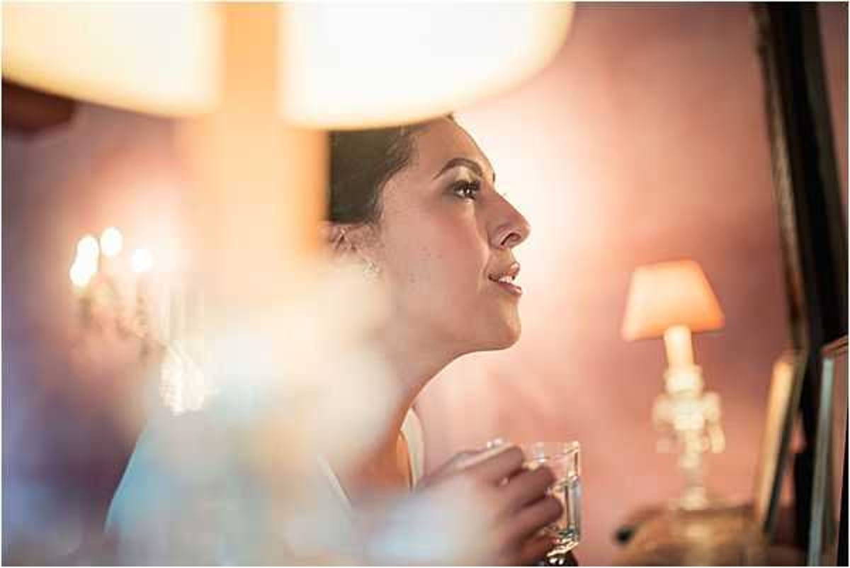 A close-up portrait of a bride during the getting-ready phase, captured in profile as she looks upward with a slight smile, holding a small glass vessel. The setting appears to be an indoor dressing room or bridal suite, warmly lit by table lamps and what appears to be a chandelier reflected in the background. The bride wears a white or ivory top and a delicate earring, with her dark hair styled up. The image has a soft, warm amber and pink tone created by the ambient lamp lighting, with intentionally blurred foreground elements — likely tulle or veil fabric — adding depth to the close-up composition.