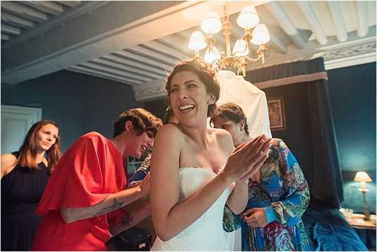 A getting-ready scene captured indoors in what appears to be a chateau room with a painted ceiling, ornate gold chandelier, and dark teal walls. The bride, wearing a strapless white dress with her hair up, laughs openly with her hands clasped together while two attendants help fasten or adjust her dress from behind — one wearing a red top and one in a blue floral robe. A fourth person in a dark dress watches from the left side of the frame. The shot is a medium close-up with warm ambient lighting from the chandelier and a bedside lamp, giving the scene a candid, joyful atmosphere.