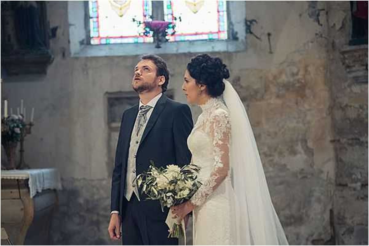 A bride and groom stand together during an indoor religious ceremony inside a historic French chapel or church. The groom wears a dark charcoal suit with a light grey waistcoat and striped tie, while the bride wears a long-sleeved ivory lace gown with a cathedral-length veil and her dark hair styled in an updo. The bride holds a loose, garden-style bouquet of white flowers and greenery with olive-toned foliage. Both are looking upward, likely toward an officiant or altar area. The church interior features aged stone walls, a colorful stained glass window, white candles on the altar, and a classic, traditional atmosphere. The shot is a medium portrait framing the couple from approximately the waist up.
