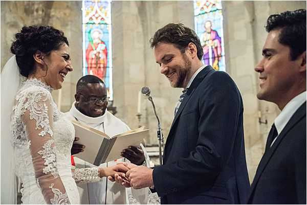 The couple is exchanging rings during an indoor church ceremony, with the groom placing the ring on the bride's finger while both smile broadly. The setting is a stone chapel with colorful stained glass windows visible in the background. The bride wears a long-sleeved white lace gown with intricate floral lace detailing and a veil, while the groom is dressed in a dark navy suit. A officiant in white liturgical robes holds an open book at a microphone stand, and a second man in a dark suit — likely a best man — stands to the right in a mid-shot composition capturing four people.